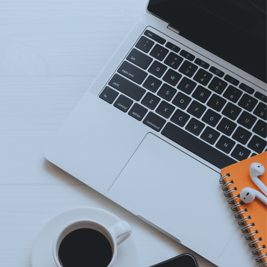 An overhead view of a laptop, a cup of coffee, an orange spiral notebook, and wireless earbuds on a white desk, representing convenient telepsychiatry from home | Blue Lotus Telepsychiatry Services, Inc.