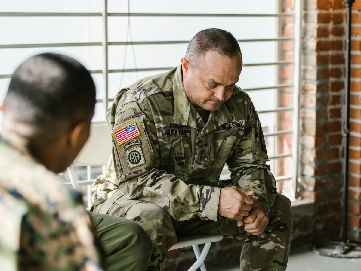 A US Army soldier in camouflage fatigues sitting indoors, looking down with a serious, pensive expression while listening to someone sitting opposite him | Blue Lotus Telepsychiatry