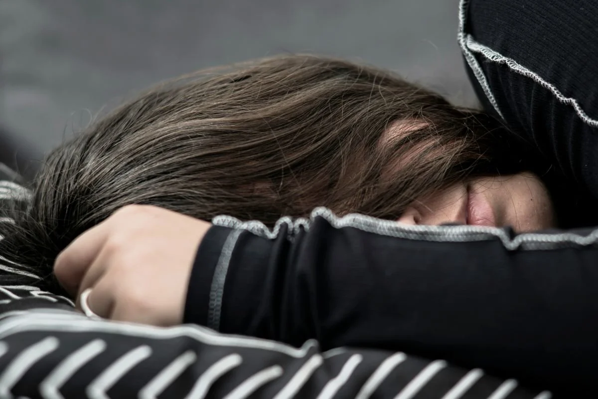 A woman lies face down, hiding her face in her hair with her arm wrapped around a striped pillow | Blue Lotus Telepsychiatry