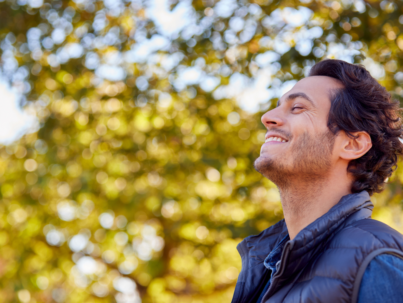 Side profile of a smiling man closing his eyes and looking up toward the sun against a background of green trees, representing trauma recovery and hope | Blue Lotus Telepsychiatry