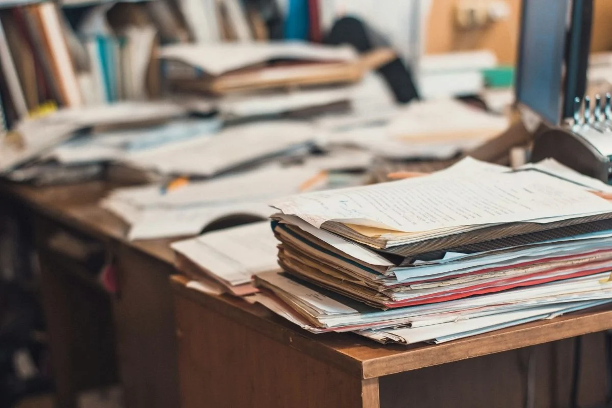 A cluttered wooden desk piled high with stacks of papers, folders, and books | Blue Lotus Telepsychiatry