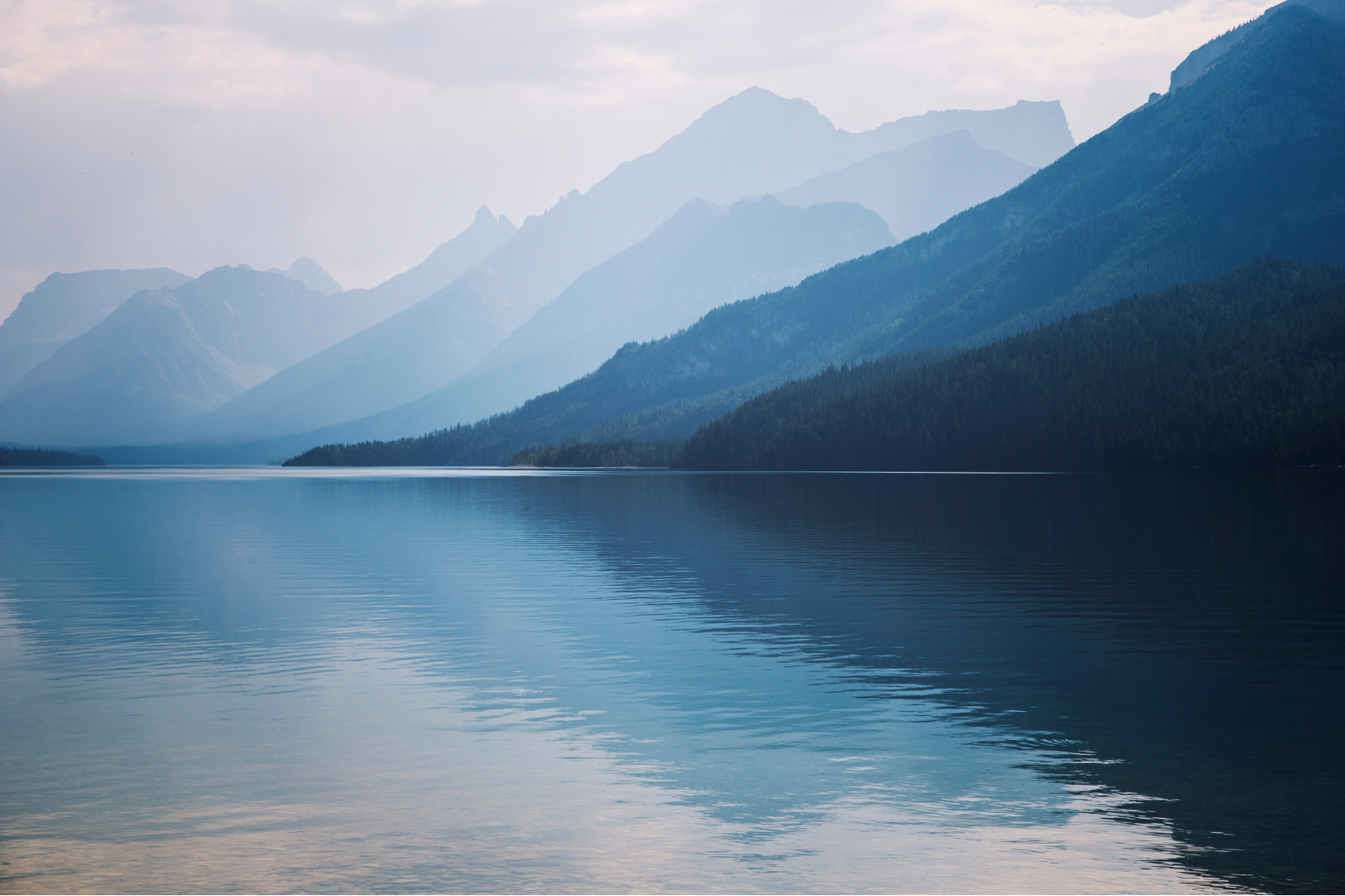 A calm lake reflecting layered blue mountain ranges fading into mist under a soft overcast sky | Blue Lotus Telepsychiatry Services, Inc.