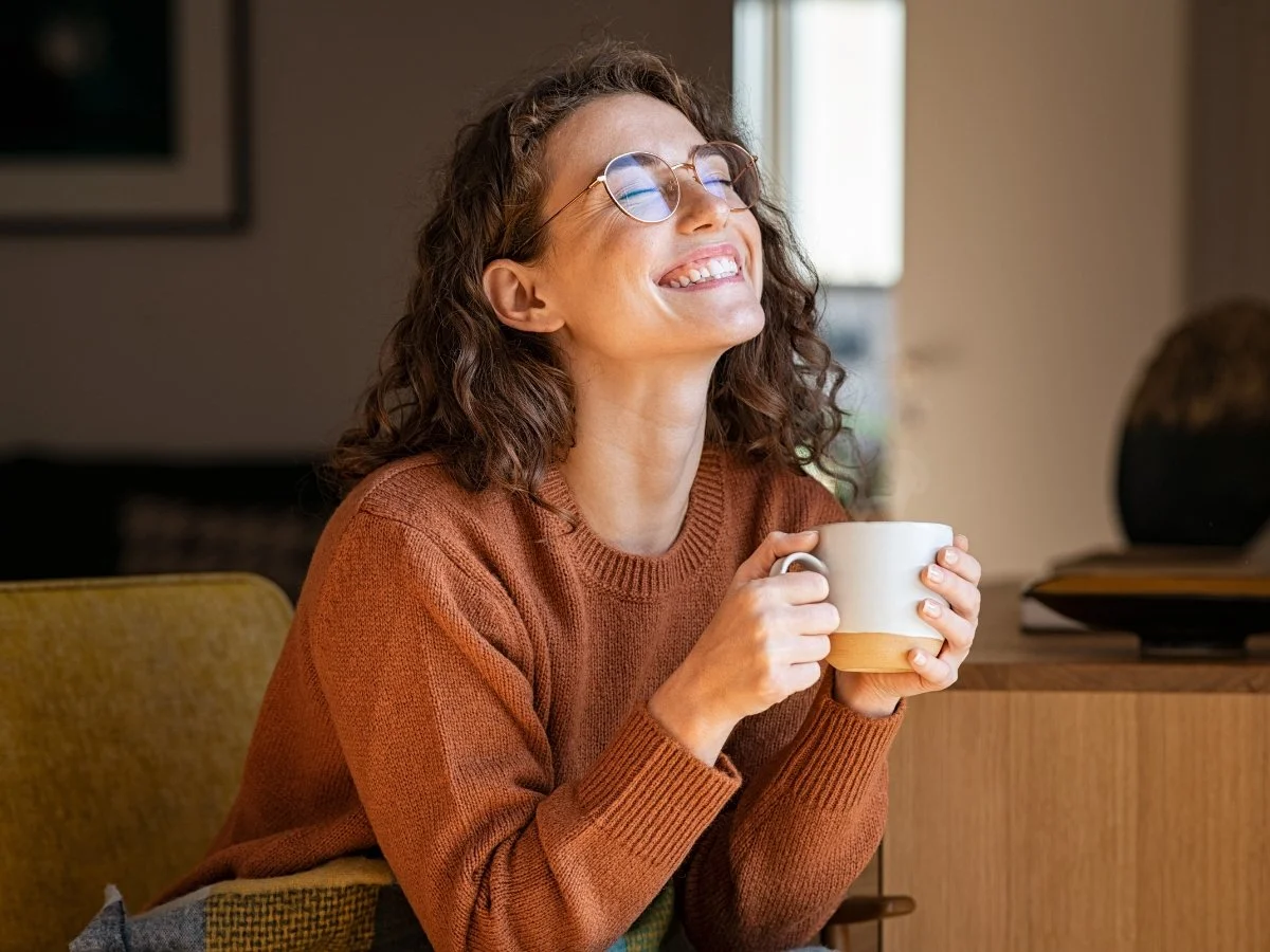 A woman wearing glasses laughs joyfully while holding a mug in a sunlit room | Blue Lotus Telepsychiatry