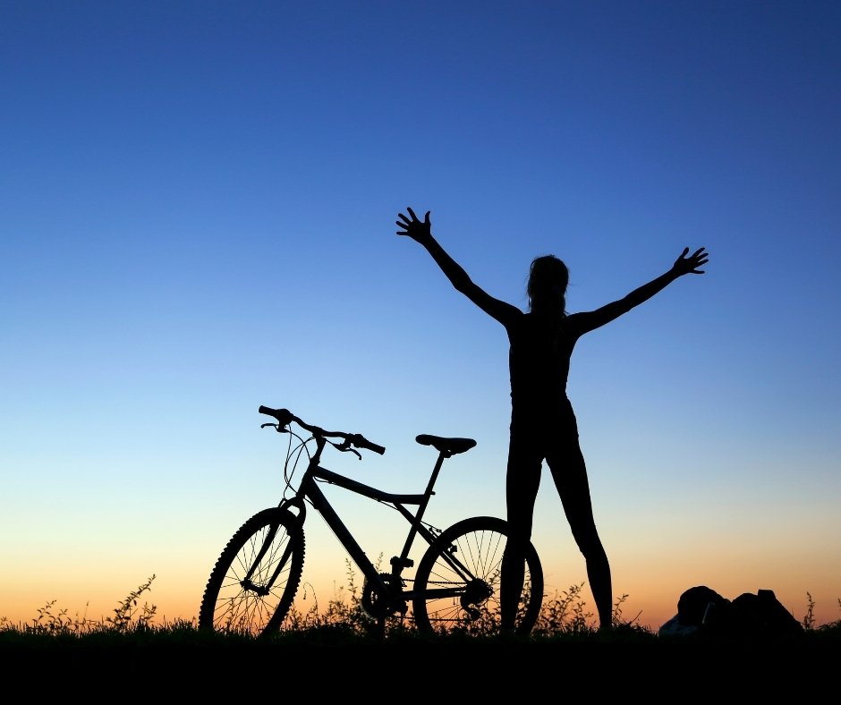 A silhouette stands beside a bicycle with arms raised toward a colorful sunset sky | Blue Lotus Telepsychiatry Services, Inc.