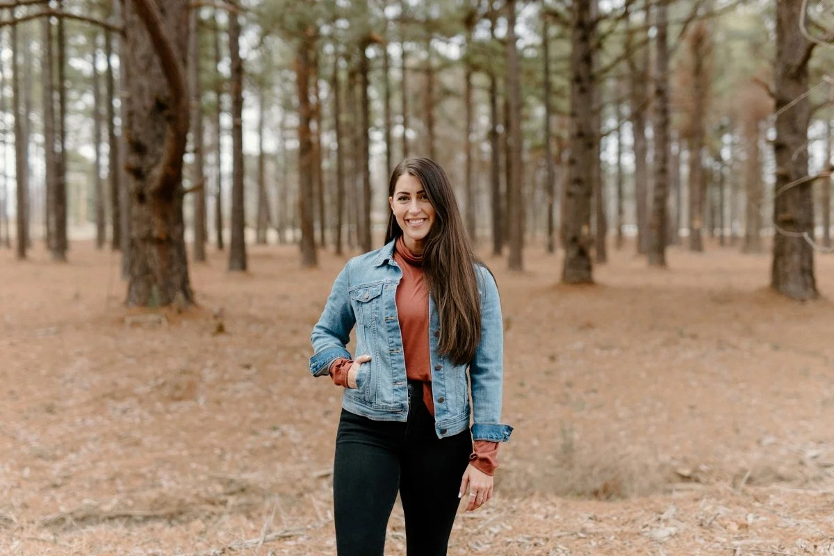 A woman in a denim jacket walks smiling through a sunlit forest | Blue Lotus Telepsychiatry