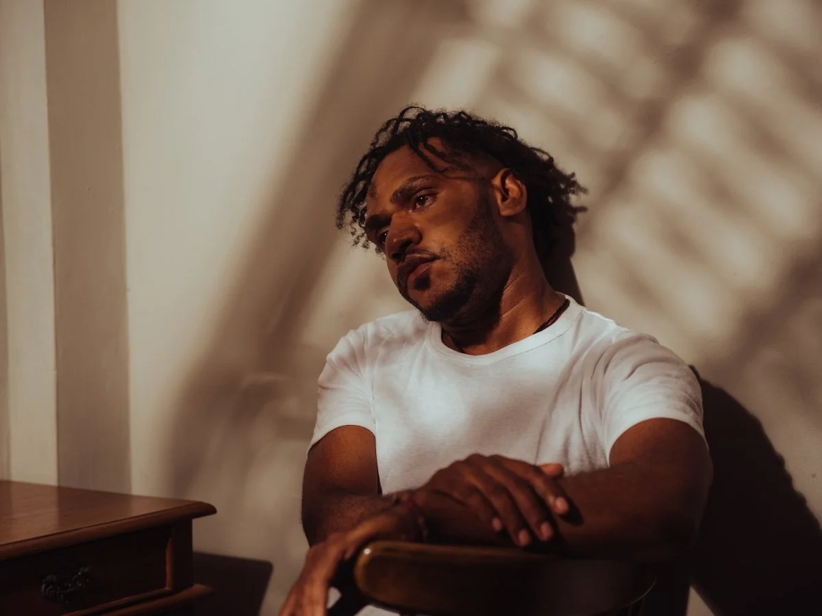 A man in a white t-shirt sitting back in a chair against a wall, leaning his head slightly to the side with a pensive, somber expression | Blue Lotus Telepsychiatry