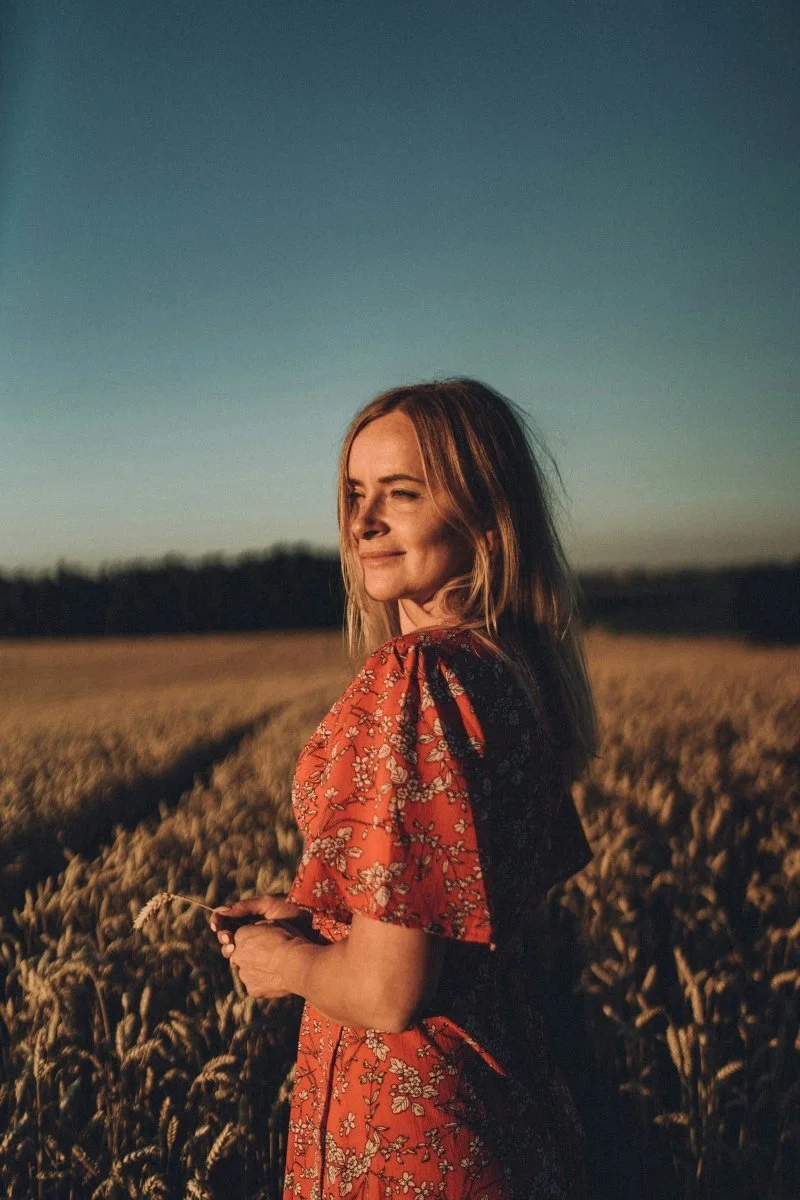 A woman in a floral dress stands in a golden wheat field at sunset, holding a stalk of wheat and smiling softly | Blue Lotus Telepsychiatry Services, Inc.