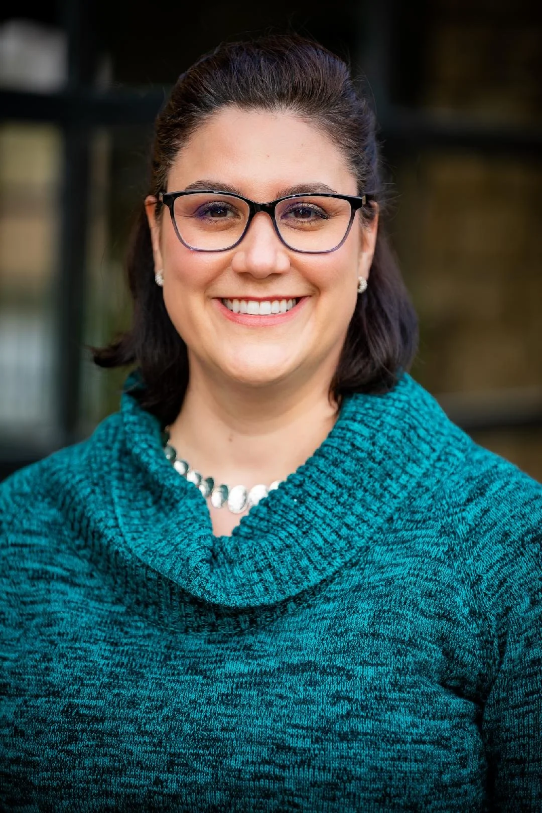 Adria Steen smiling and wearing glasses and a teal cowl neck sweater, photographed outdoors with a blurred building background | Blue Lotus Telepsychiatry Services, Inc.