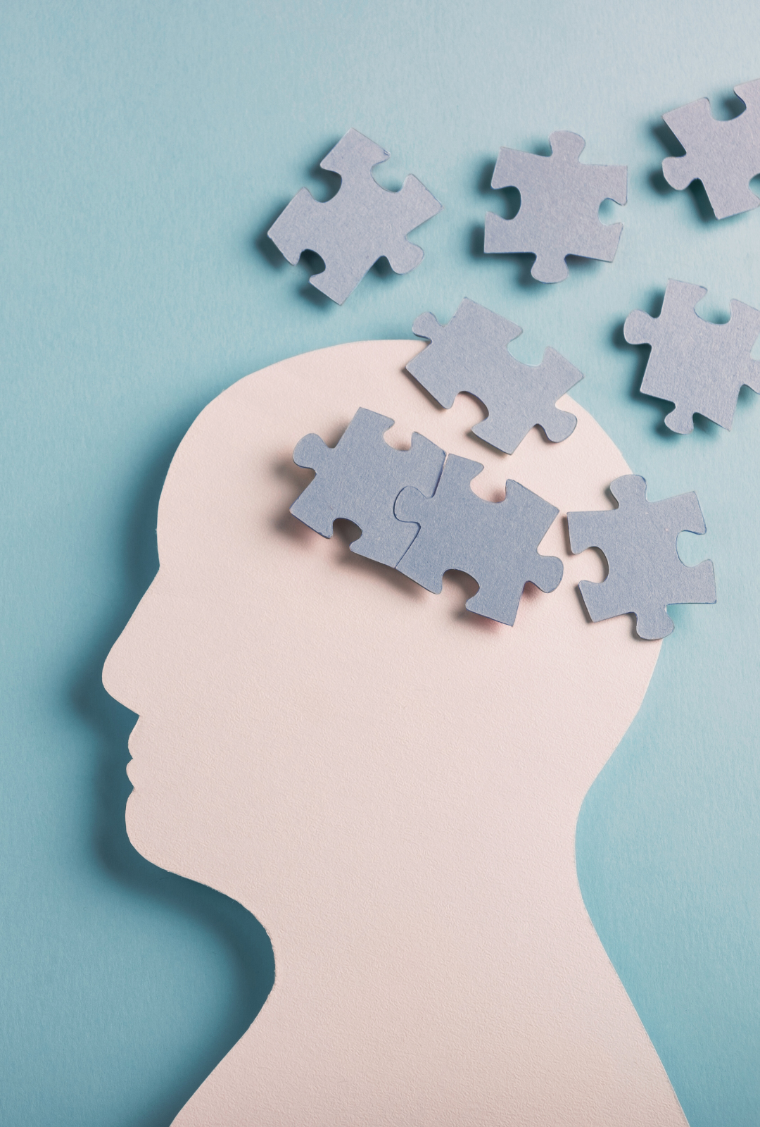 A paper cutout of a head silhouette surrounded by scattered puzzle pieces on a light blue background, symbolizing mental health and pieces coming together | Blue Lotus Telepsychiatry Services, Inc.