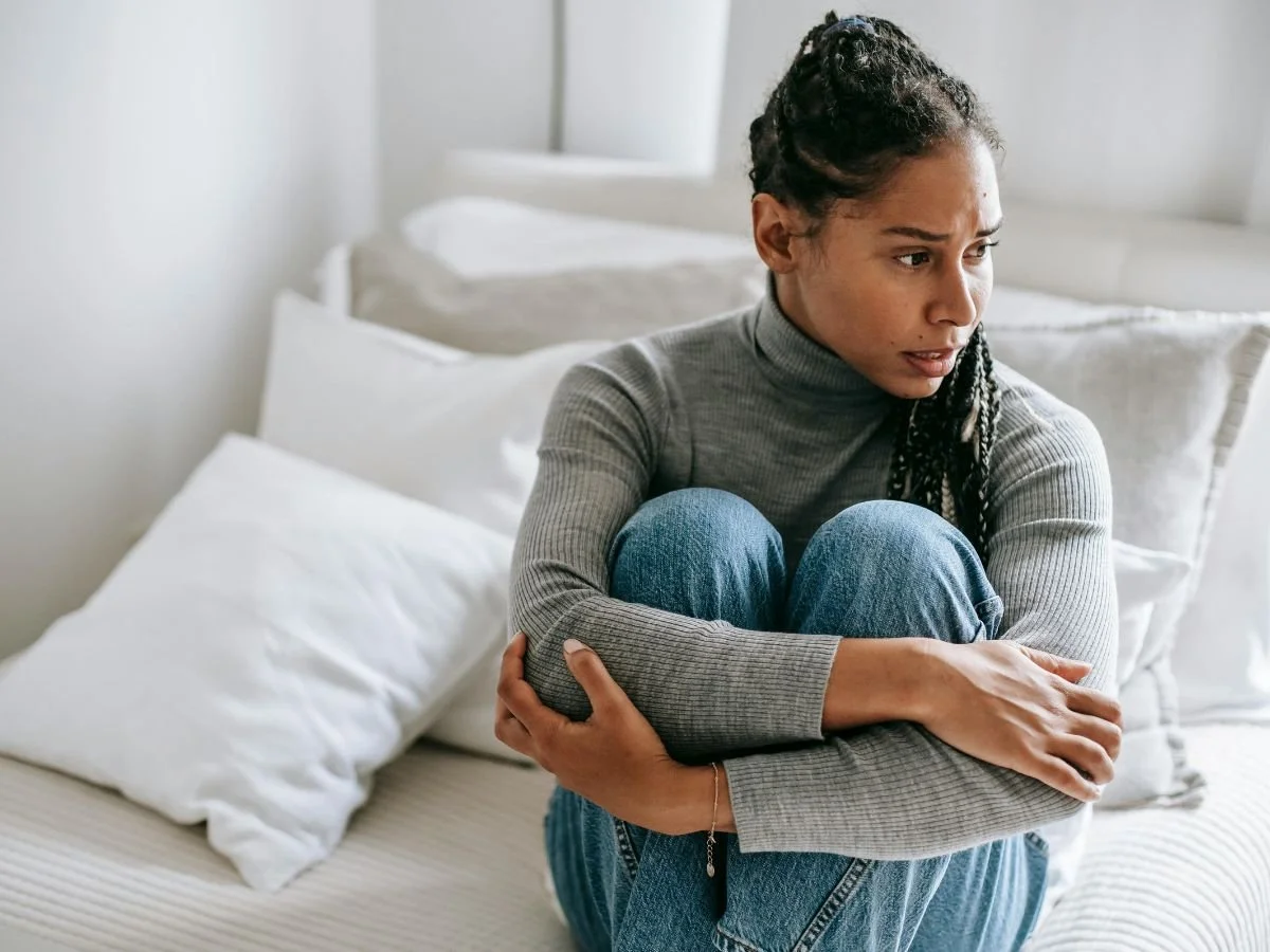 A woman sits on a couch hugging her knees to her chest, her brow furrowed with a worried expression as she looks off to the side | Blue Lotus Telepsychiatry