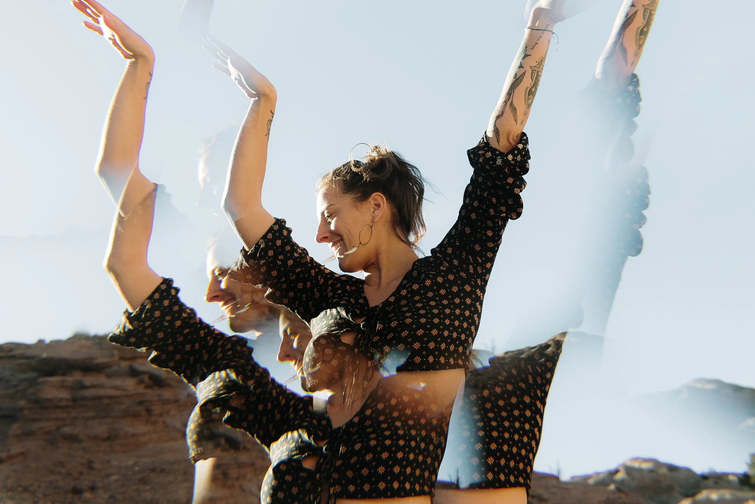 Two women in black patterned outfits performing a synchronized dance or yoga pose outdoors with arms raised, against a clear sky and rocky terrain.