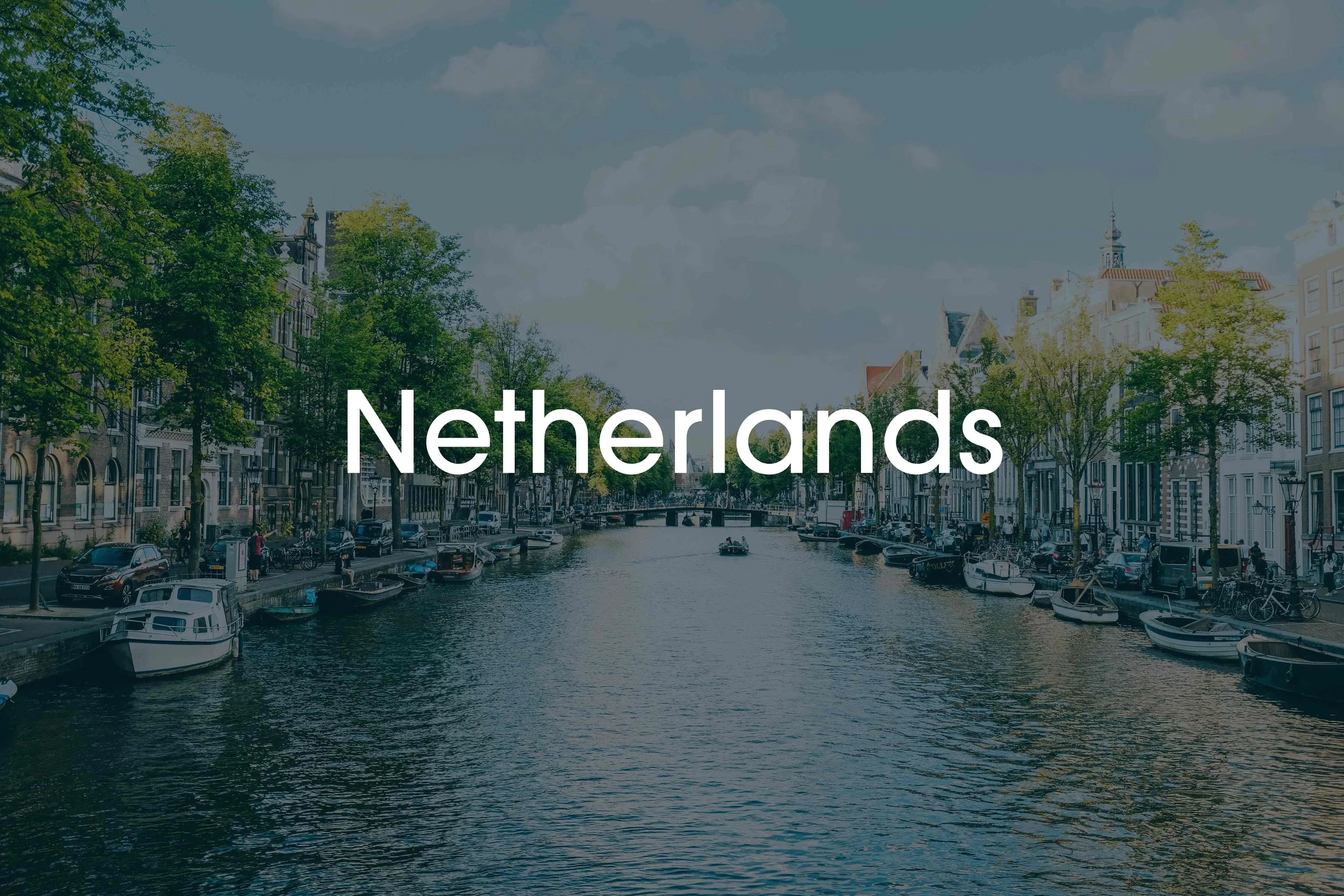 A canal in the Netherlands with trees lining both sides and boats docked along the edges, under a partly cloudy sky.
