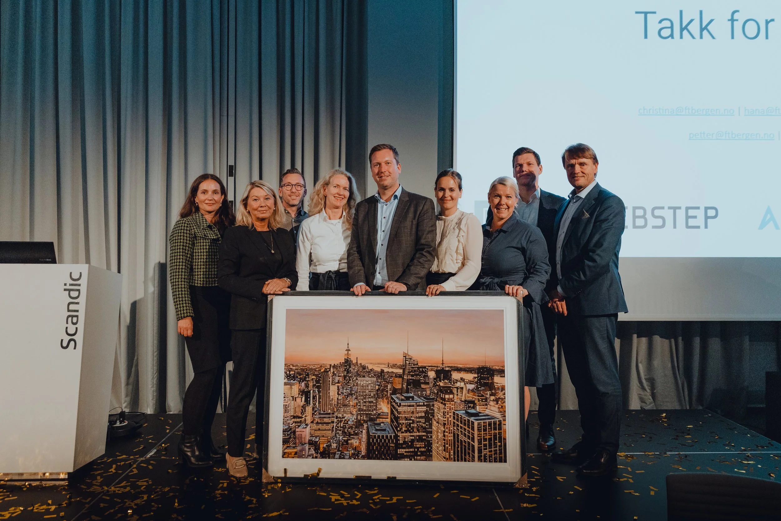 Group of ten people standing on a stage behind a large framed picture of a city skyline, with a presentation screen and a white panel marked 'Scandic' in the background, at a corporate event or award ceremony.