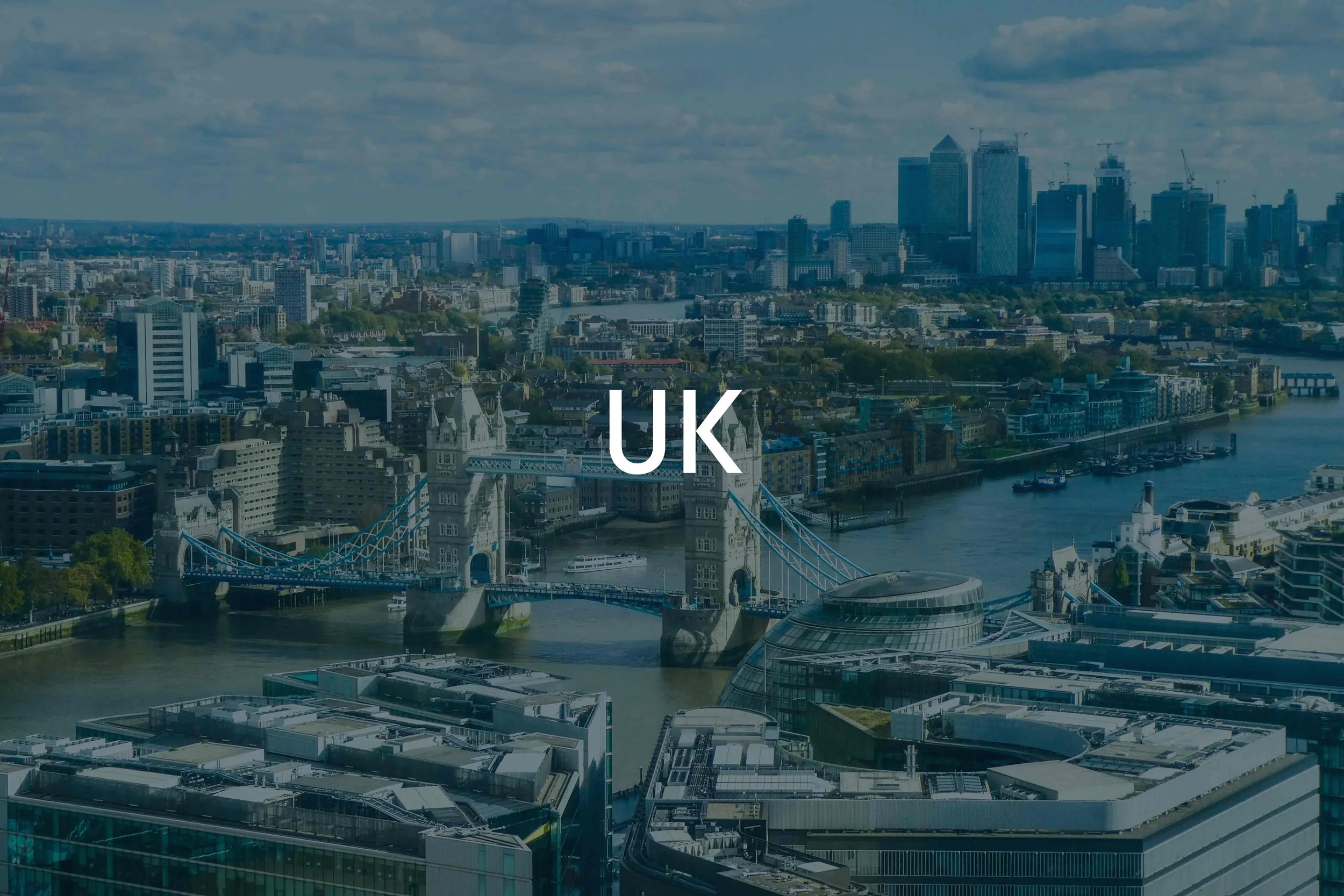 An aerial view of London's cityscape featuring Tower Bridge over the River Thames, with modern skyscrapers and historic buildings in the background, under a cloudy sky.