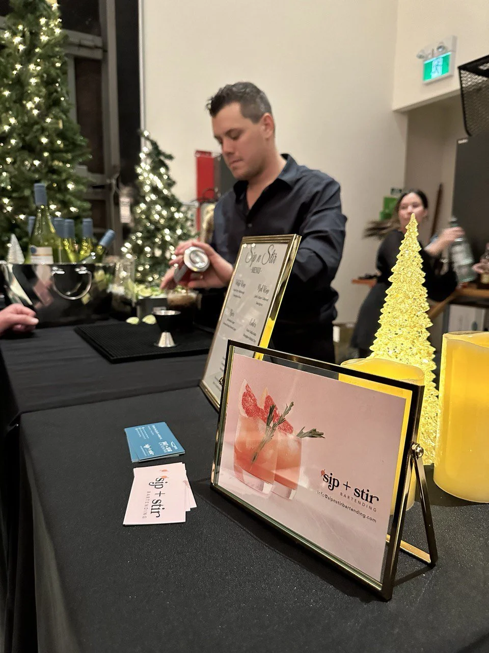 A bartender preparing drinks at a cocktail event, with a menu labeled "sip + stir." A framed picture of a cocktail, business cards, and decorative lights are on a black tablecloth. Christmas trees with lights are in the background.