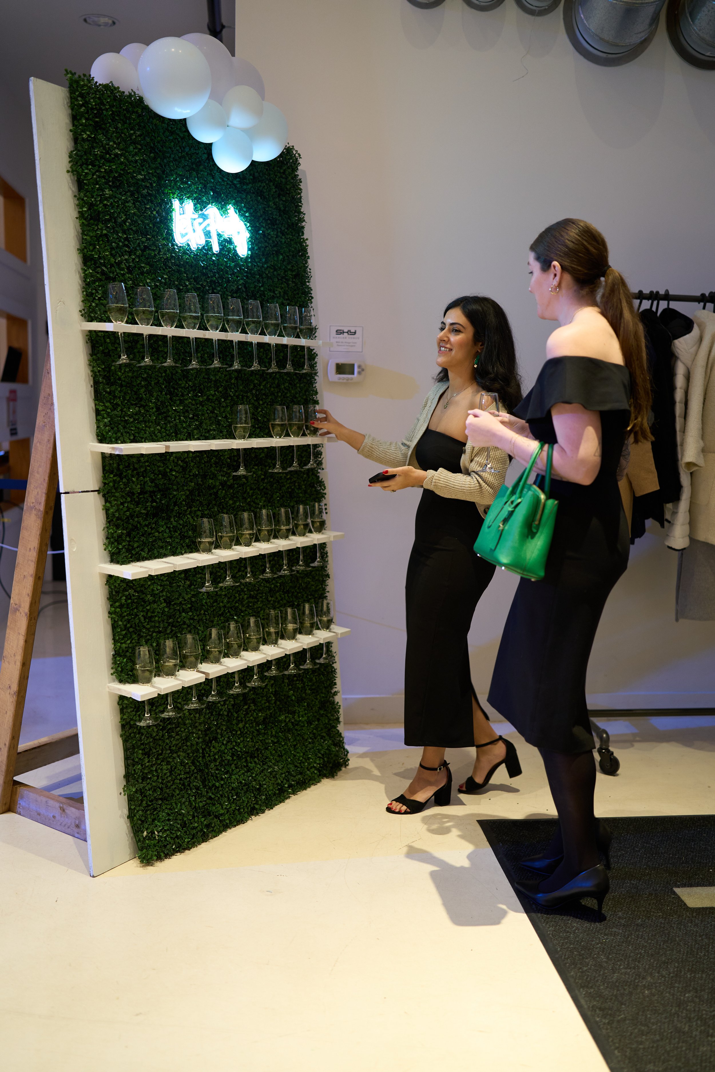 Two women in formal dresses standing near a decorative wall with shelves holding champagne glasses and illuminated signage. One woman is holding a green handbag.