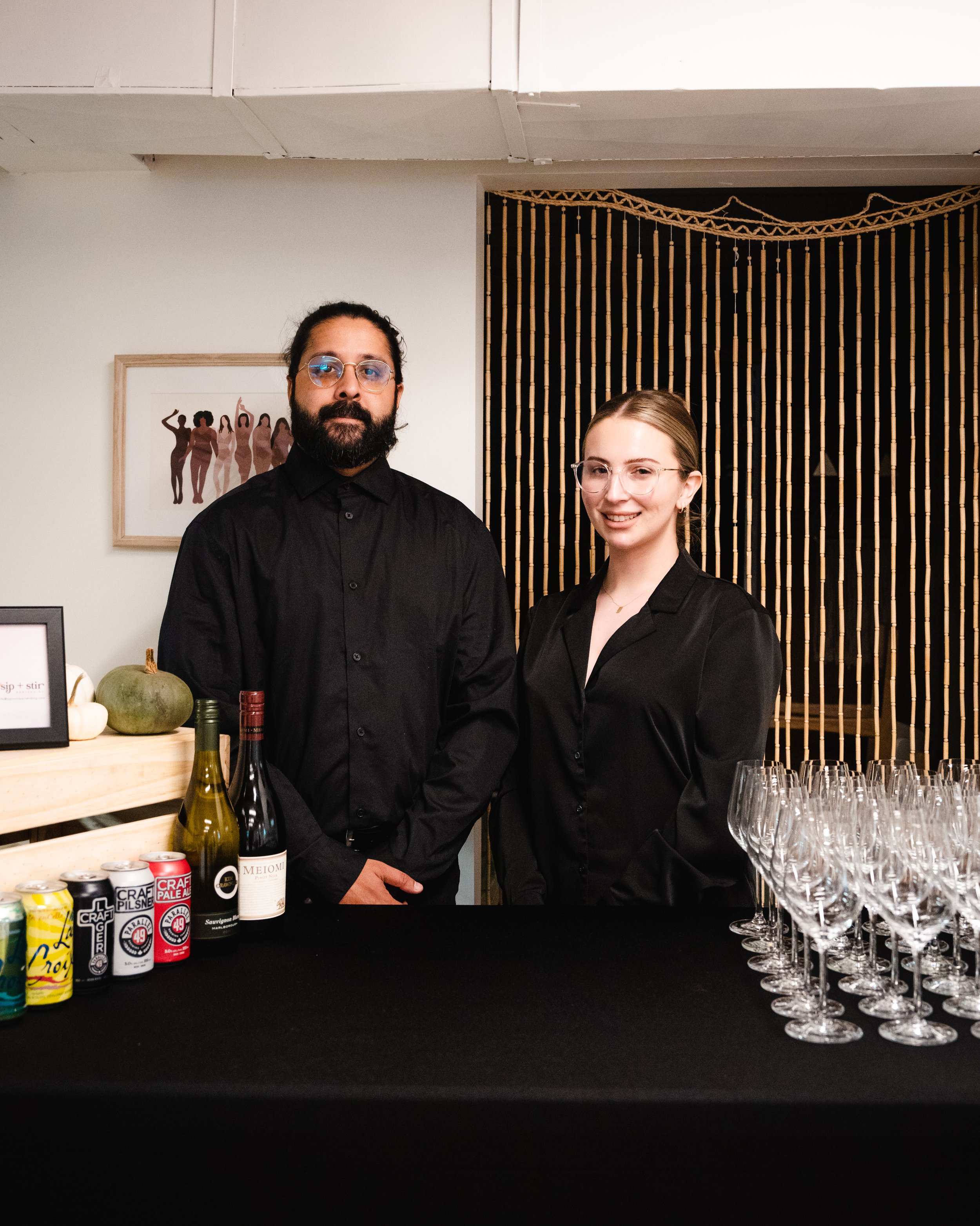 Two people in black attire standing behind a black table with drink cans, wine bottles, and a row of empty wine glasses. A wooden crate with a framed picture and a small pumpkin is to the left. A decorative wall art is visible in the background.