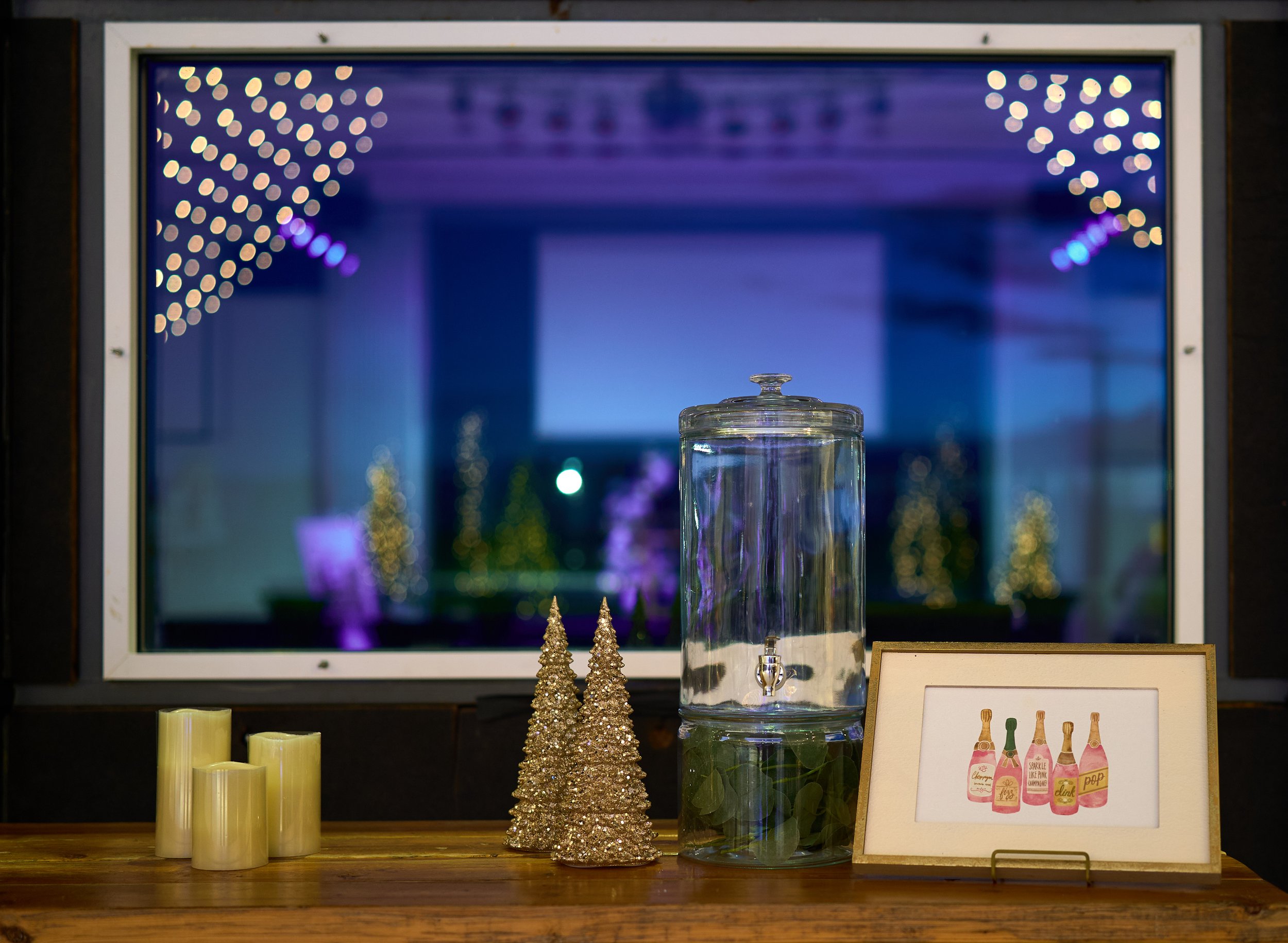 Table setup with decorative items including gold Christmas tree figurines, a glass beverage dispenser, candles, and a framed watercolor painting of champagne bottles. Blurred background with lights and trees.