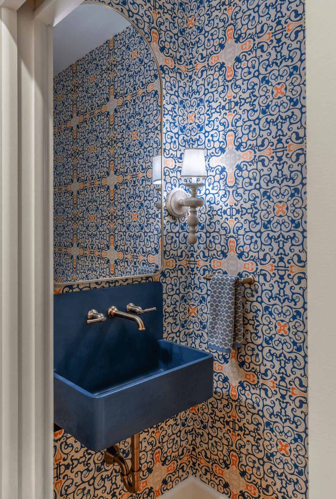 A small bathroom with patterned blue, orange, and white wallpaper, a dark blue sink, a wall-mounted lamp with a white shade, a mirror, and a towel rack with a patterned towel.