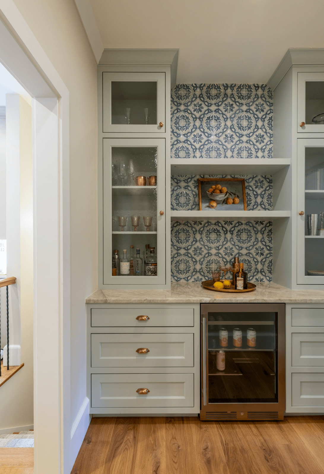Kitchen cabinet with glass doors, blue and white patterned backsplash, countertop with a tray, bottles, lemons, and a small refrigerator below.