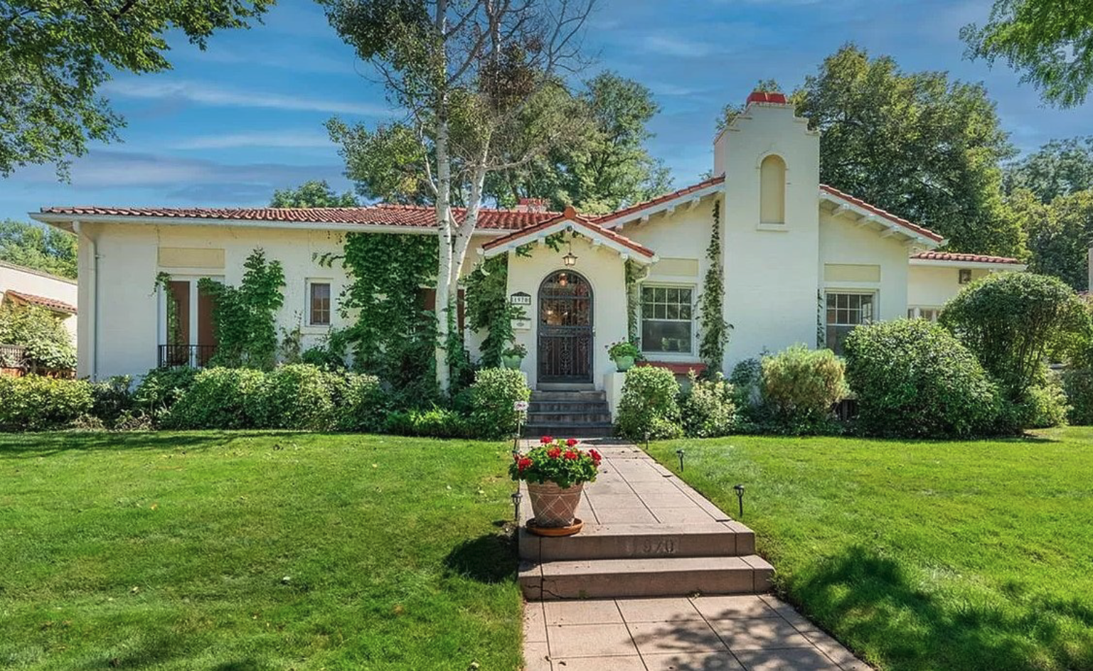 The Sinclair Home Remodel. A white house with a red tile roof, surrounded by green trees and shrubs, with a paved walkway leading to the front door and a large potted plant with red flowers on the steps.