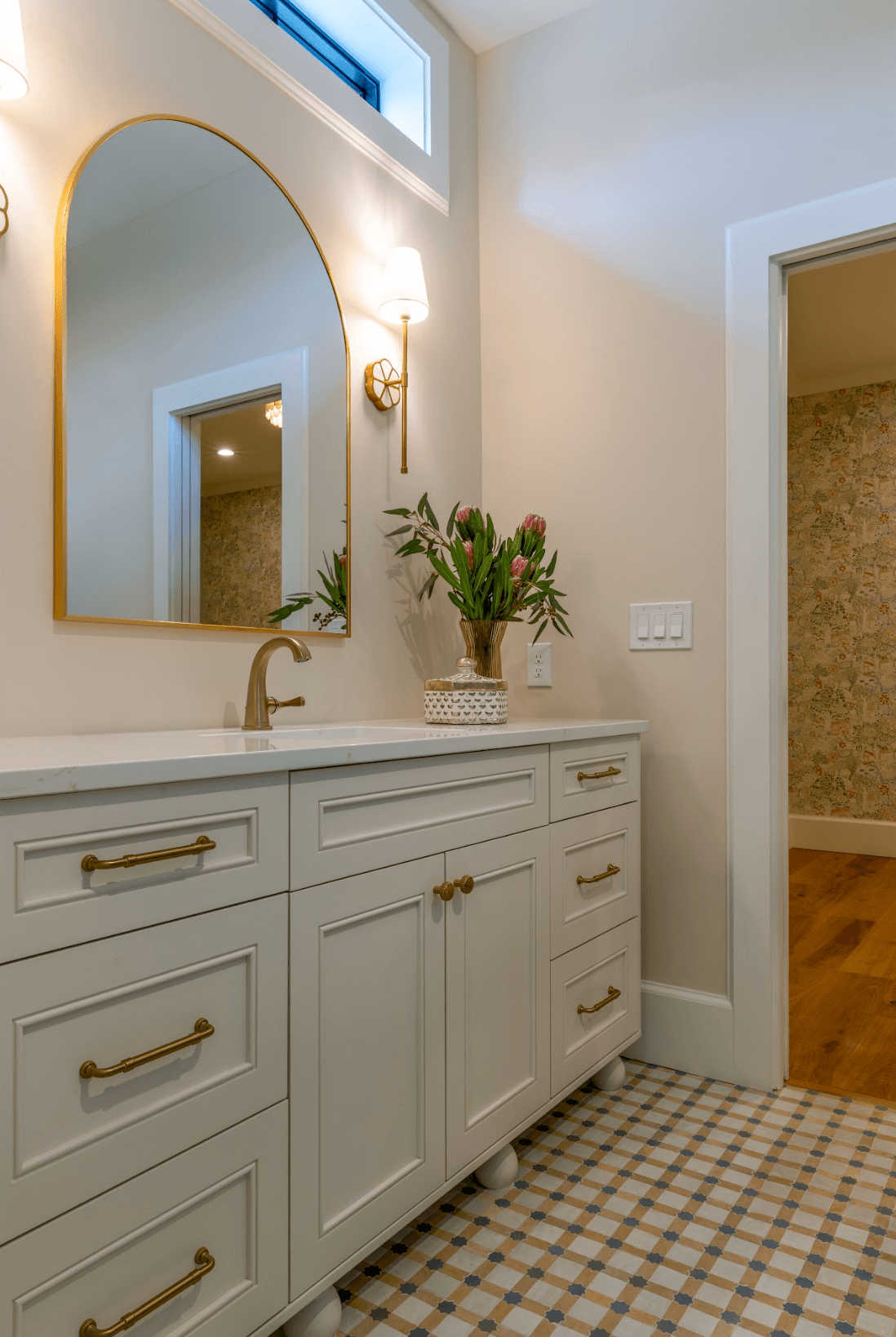 Bathroom vanity with a white cabinet, gold handles, a gold faucet, a large mirror with a gold frame, a wall-mounted light with a shade, a vase with pink flowers, and patterned tile flooring.
