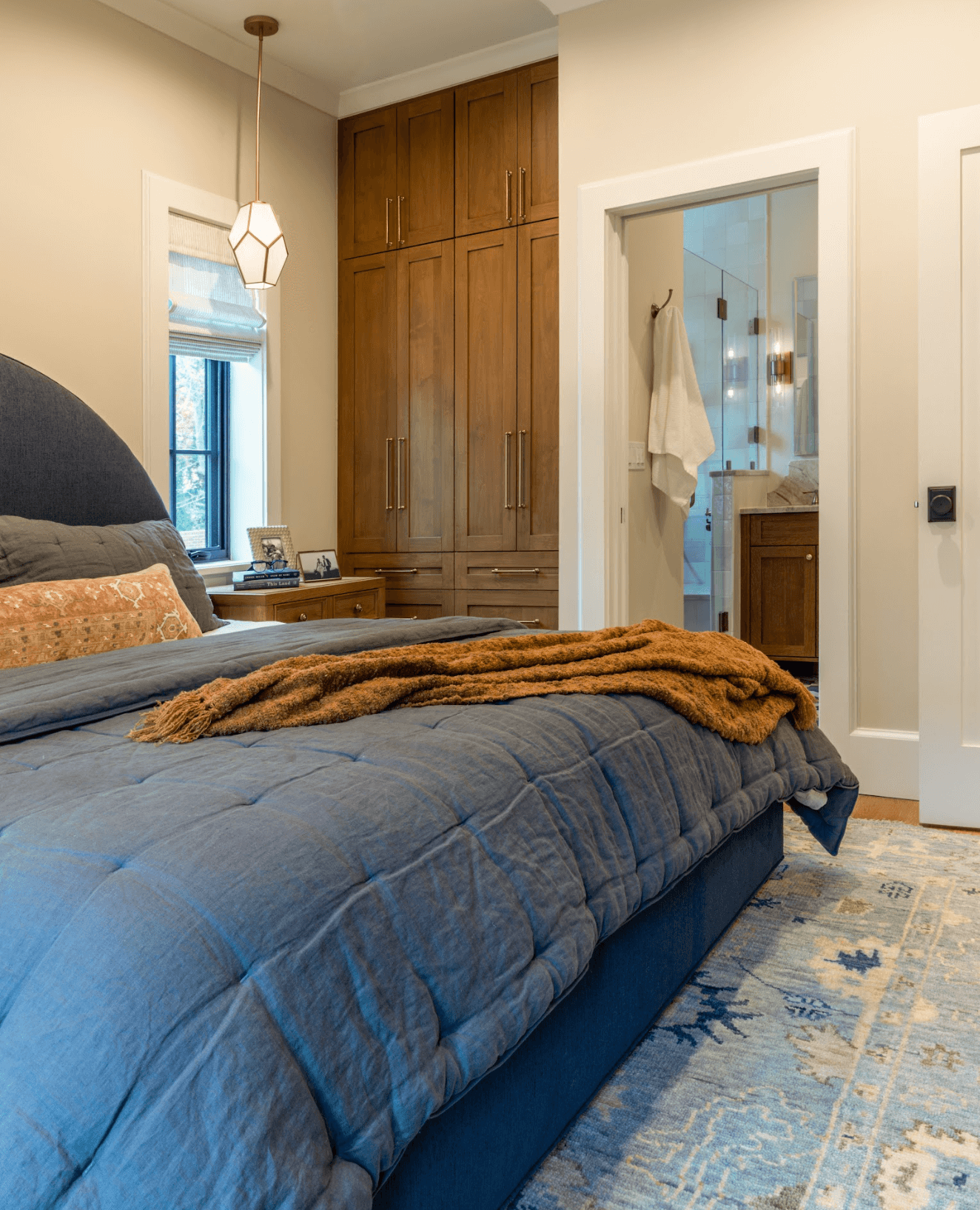 A cozy bedroom with a bed featuring a dark blue quilt, pillows, and a brown throw blanket. The room includes a wooden nightstand with framed photos, a window with a blind, a large wooden closet, and a view of a bathroom through an open door. A geomet
