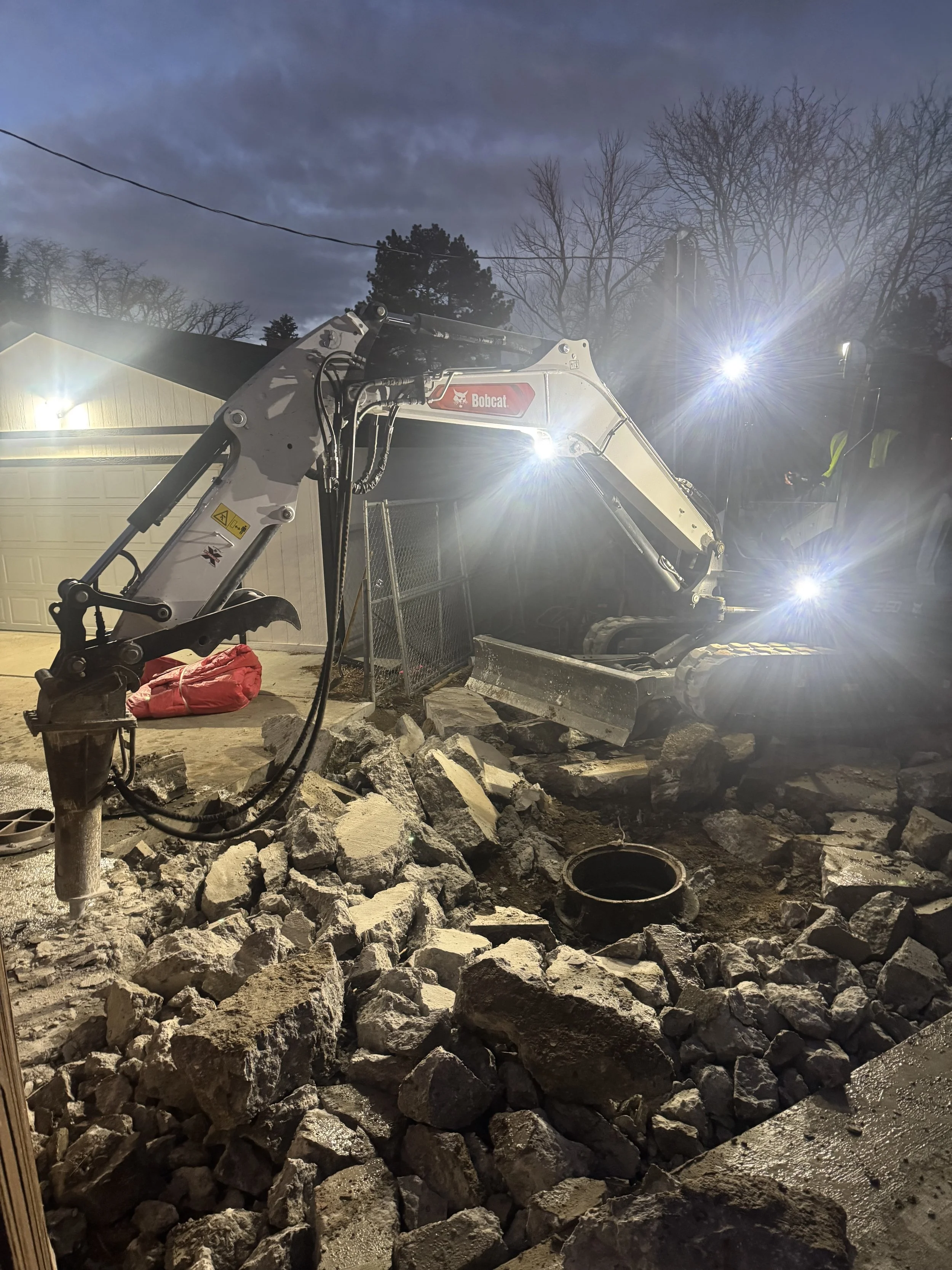 The Elizabeth Victorian-style home addition. Mid-project construction site at dusk with a Bobcat excavator moving large rocks and debris, illuminated by bright work lights.