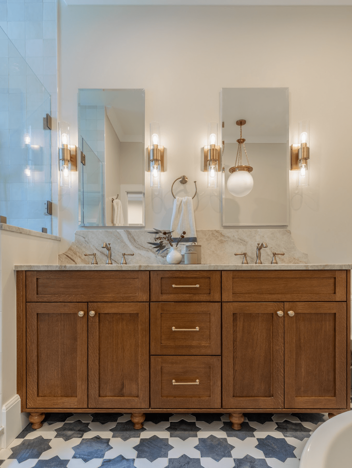 A modern bathroom vanity with a wooden cabinet, marble countertop, two faucets, and two mirrors with wall-mounted lights on either side. A vase with decorative branches is on the countertop. The floor has a black and white geometric patterned tile.