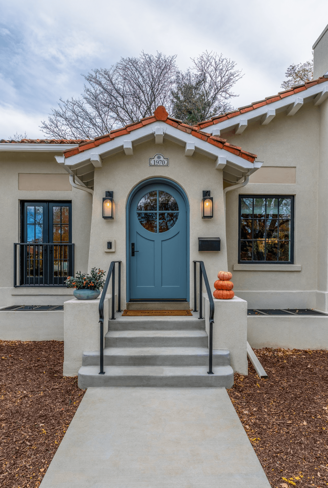 Front entrance of a house with a blue door, black railing stairs, and fall decor including pumpkins and a plant. The house has beige walls, two lantern lights, and a sign with the year 1970 above the door. There are trees with no leaves and a cloudy 
