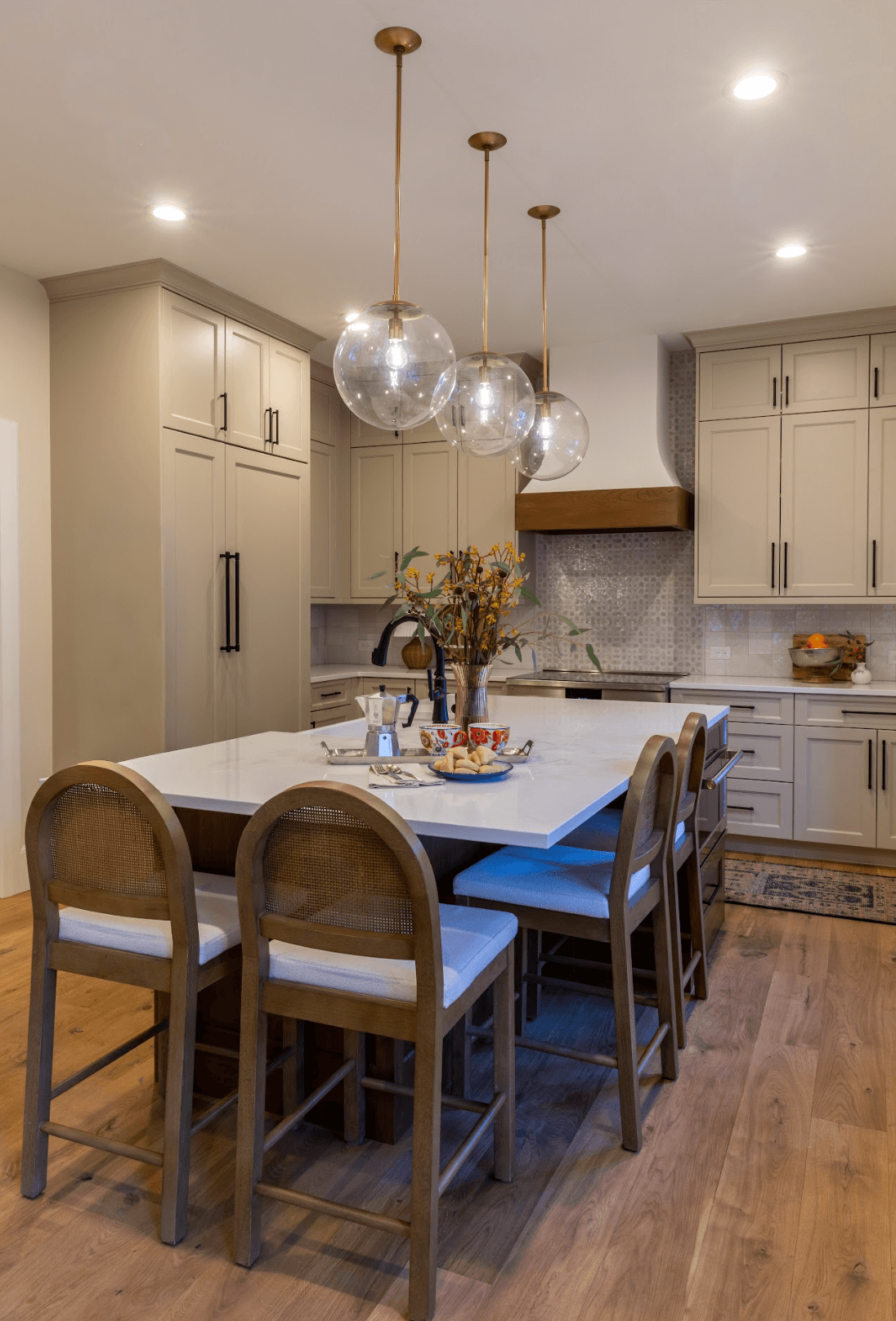 Modern kitchen with beige cabinets, a white island, wooden bar stools, and pendant lights with glass globes.