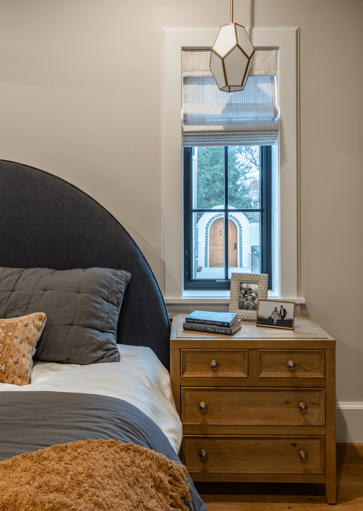 A cozy bedroom corner with a dark upholstered bed, gray pillows, a wooden nightstand with books and framed photos, a window with a Roman shade, and a hanging pendant light.