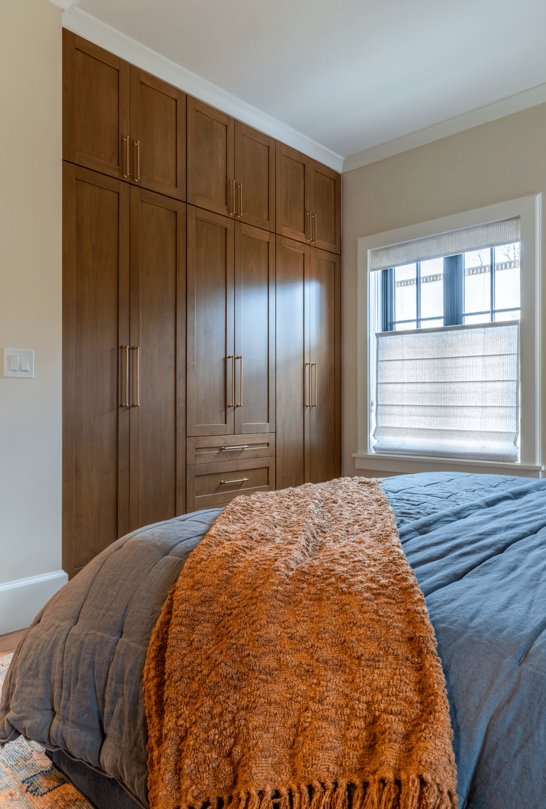 A bedroom with a large wooden wardrobe beside a window covered with a white shade. The bed has a dark blue comforter and an orange textured throw blanket at the foot.