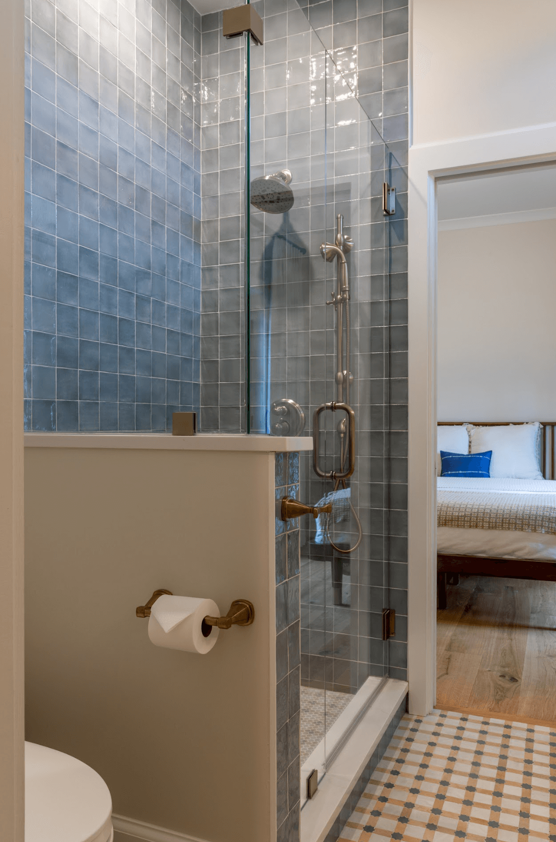 Bathroom shower with glass door, blue tiles, and brass fixtures. A toilet with a paper roll holder nearby.