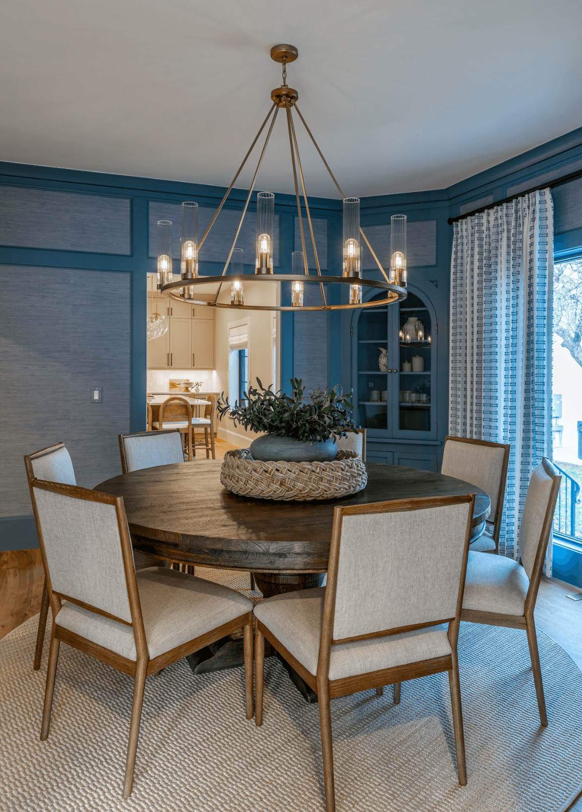A round wooden dining table with six beige upholstered chairs, a decorative centerpiece with greenery, a modern chandelier hanging above, blue walls, and windows with blue and white checked curtains.