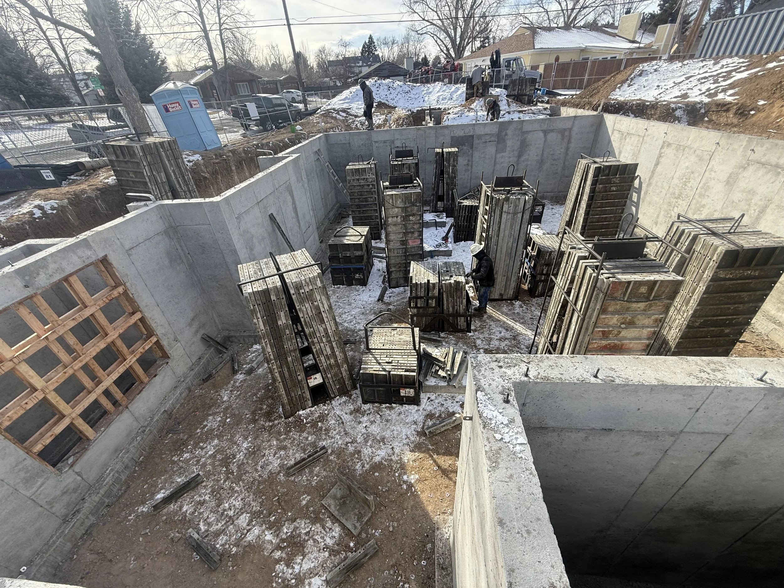The Elizabeth Victorian-style home addition. Mid-project construction construction site with concrete foundation, metal framing, and workers installing support structures, snowy ground, and residential area in background.