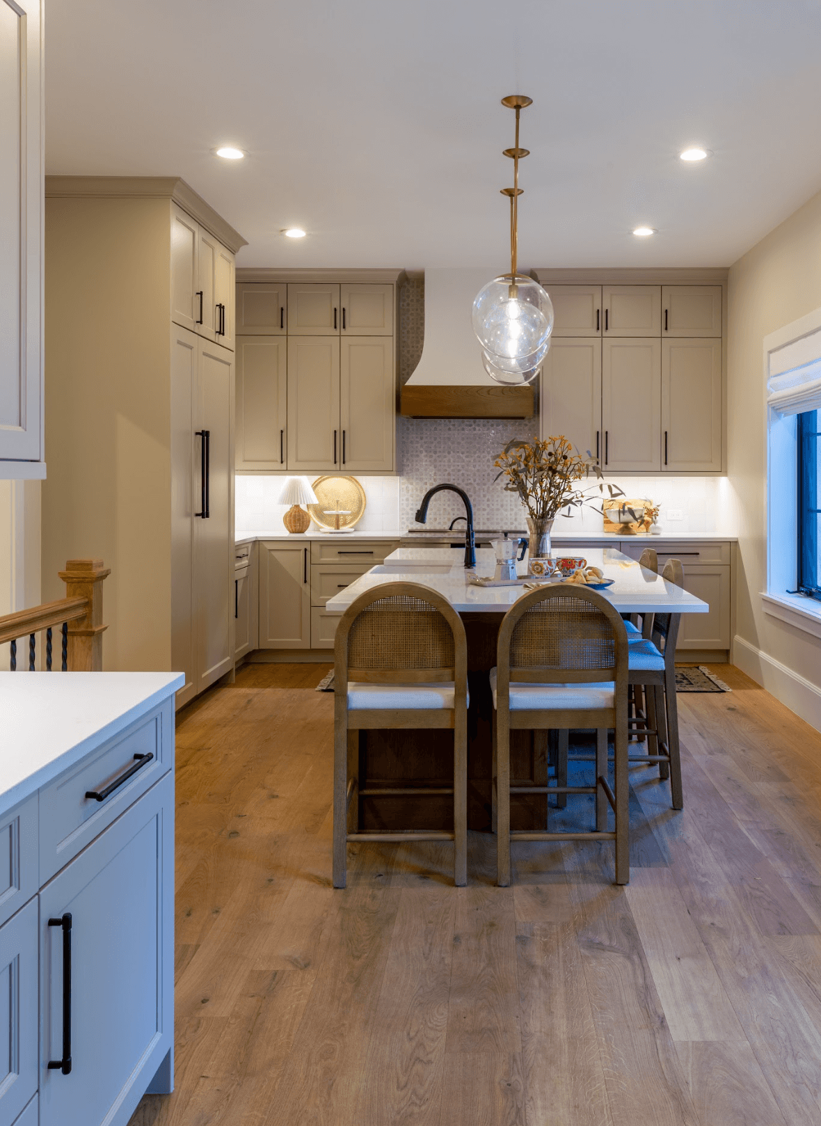 A modern kitchen with light gray cabinets, a kitchen island with barstools, a pendant light, and a window with blinds.