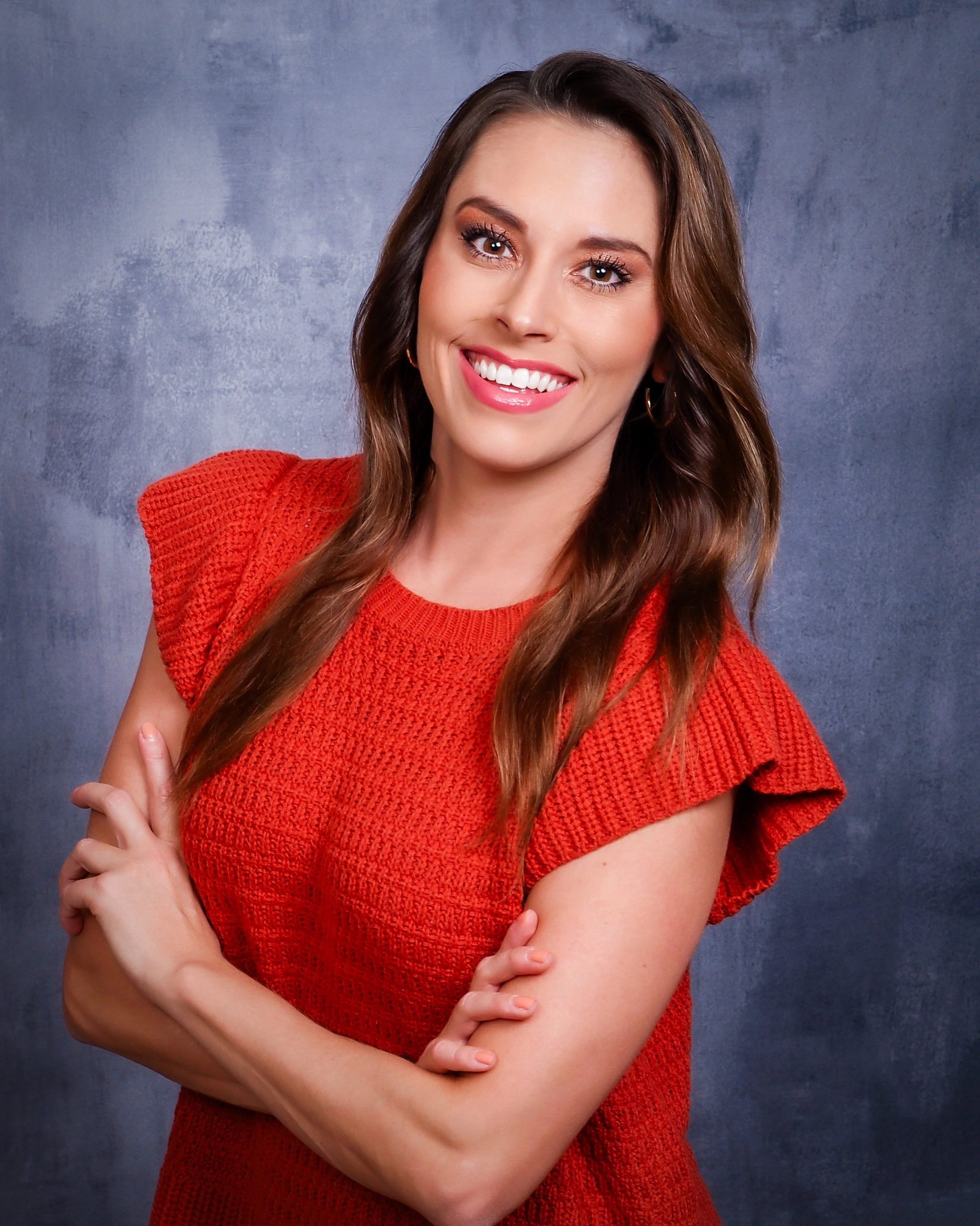A smiling woman with long brown hair wearing a red textured top with ruffled sleeves, standing against a mottled gray background.