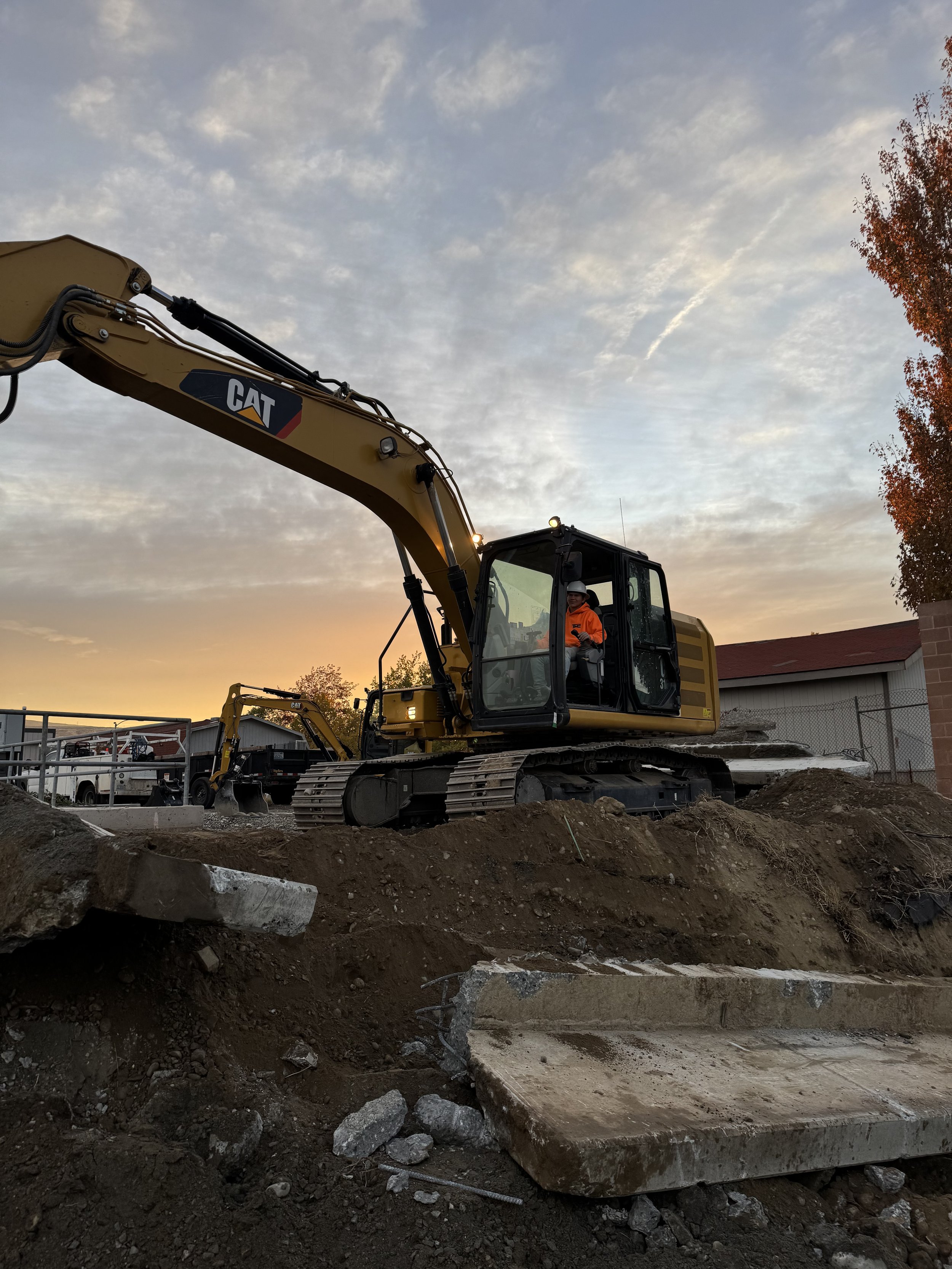 A construction site at sunset with a yellow CAT excavator. The operator is sitting inside the cab wearing an orange safety jacket and a helmet. Dirt, rocks, and a concrete slab are in the foreground, with a tree with autumn leaves and a building in the background.