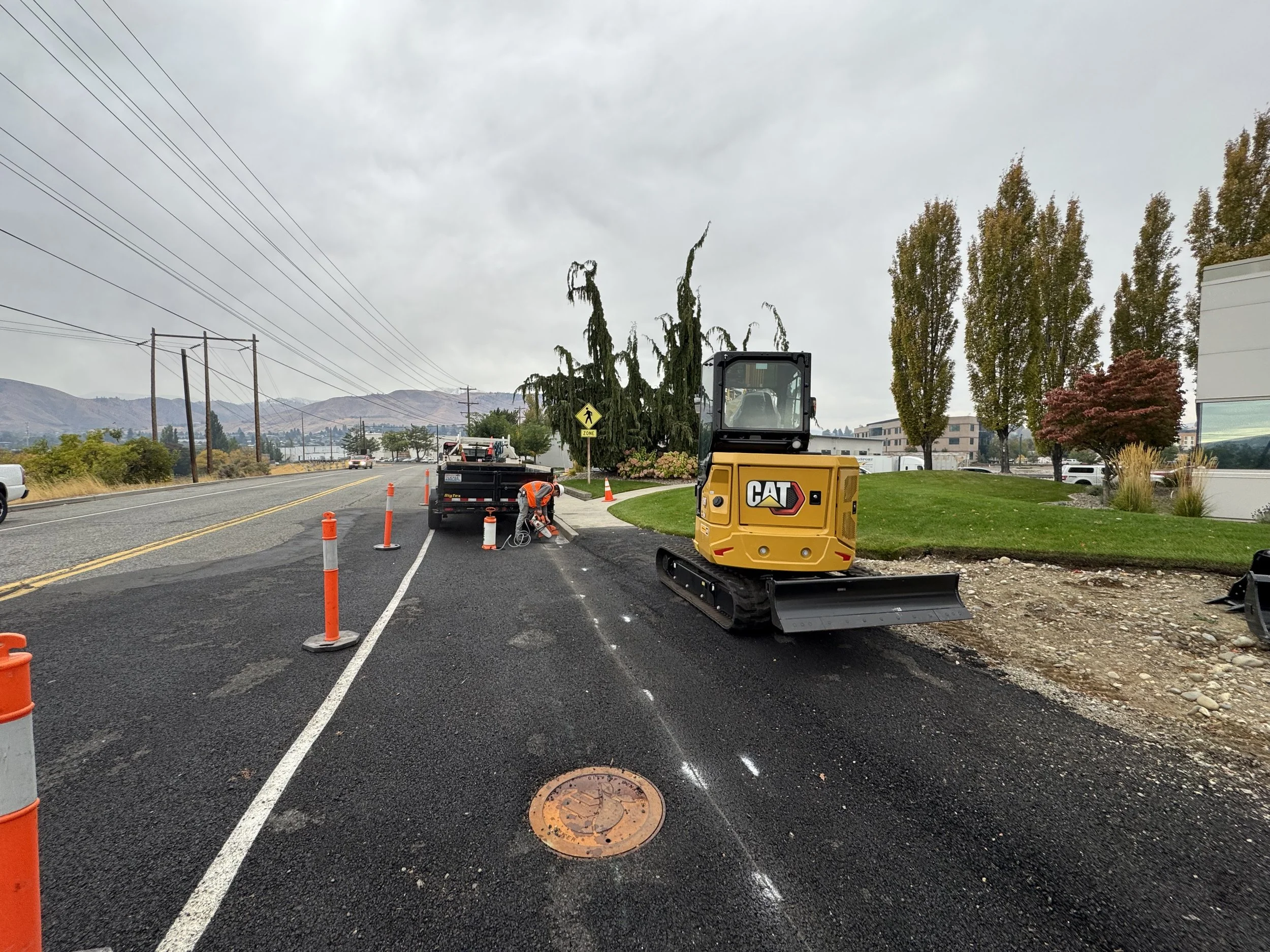 Workers repairing a section of road with orange traffic cones, a yellow CAT excavator, and a black dump trailer along a road with trees, a building, and mountains in the background under cloudy skies.