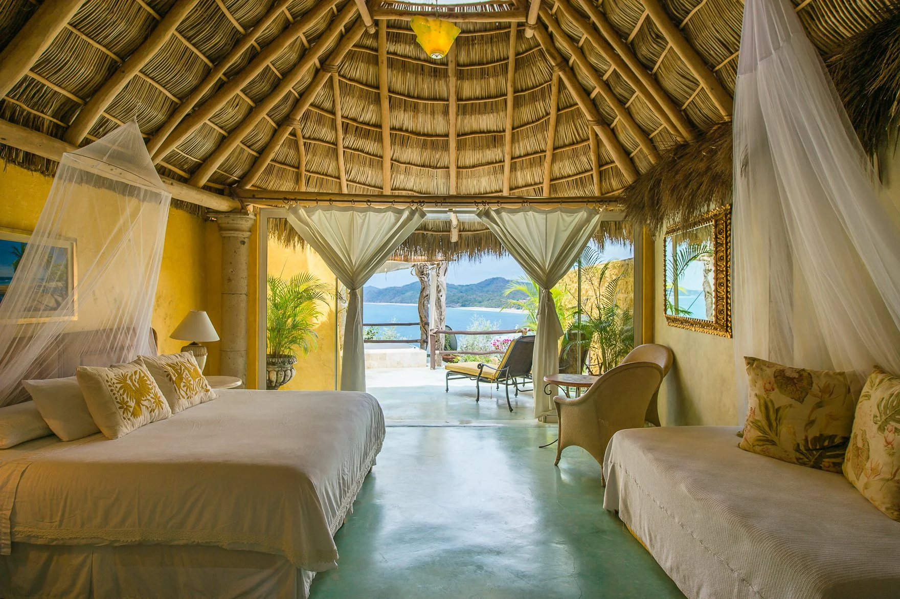 A tropical bedroom with a thatched roof, view of a balcony, lake, and mountains through open curtains, with beds, chairs, and potted plants inside.