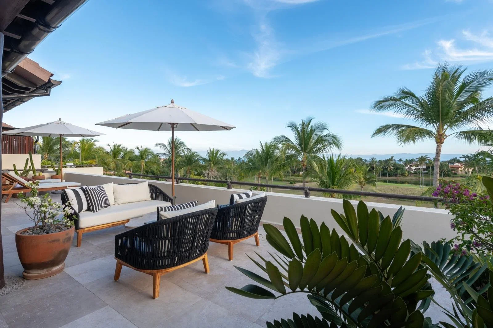 A spacious outdoor patio area with white sofas, black and white striped pillows, potted plants, and large white umbrellas. The view overlooks a lush landscape with numerous palm trees and a distant body of water under a clear blue sky.