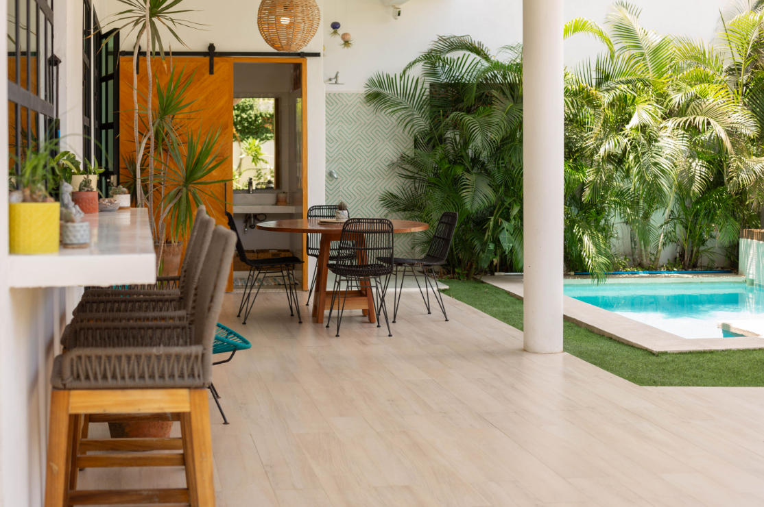 Indoor patio with dining table, black chairs, potted plants, pool, and lush green tropical foliage outside.