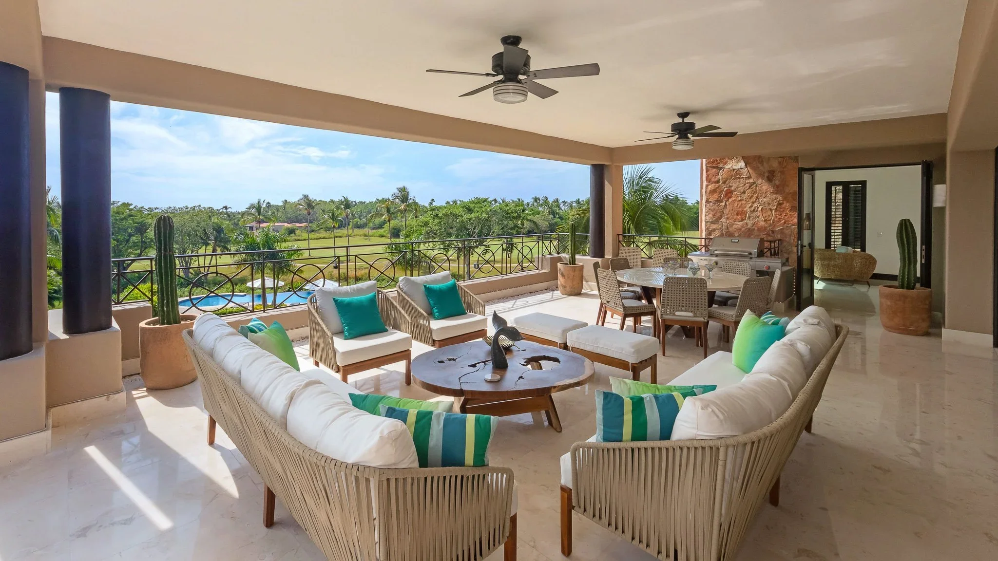 Covered outdoor patio with seating area including a semi-circular white sofa, chairs, and colorful cushions. There is a round coffee table, a dining table with chairs, a gas grill, potted cacti, and tropical scenery in the background.