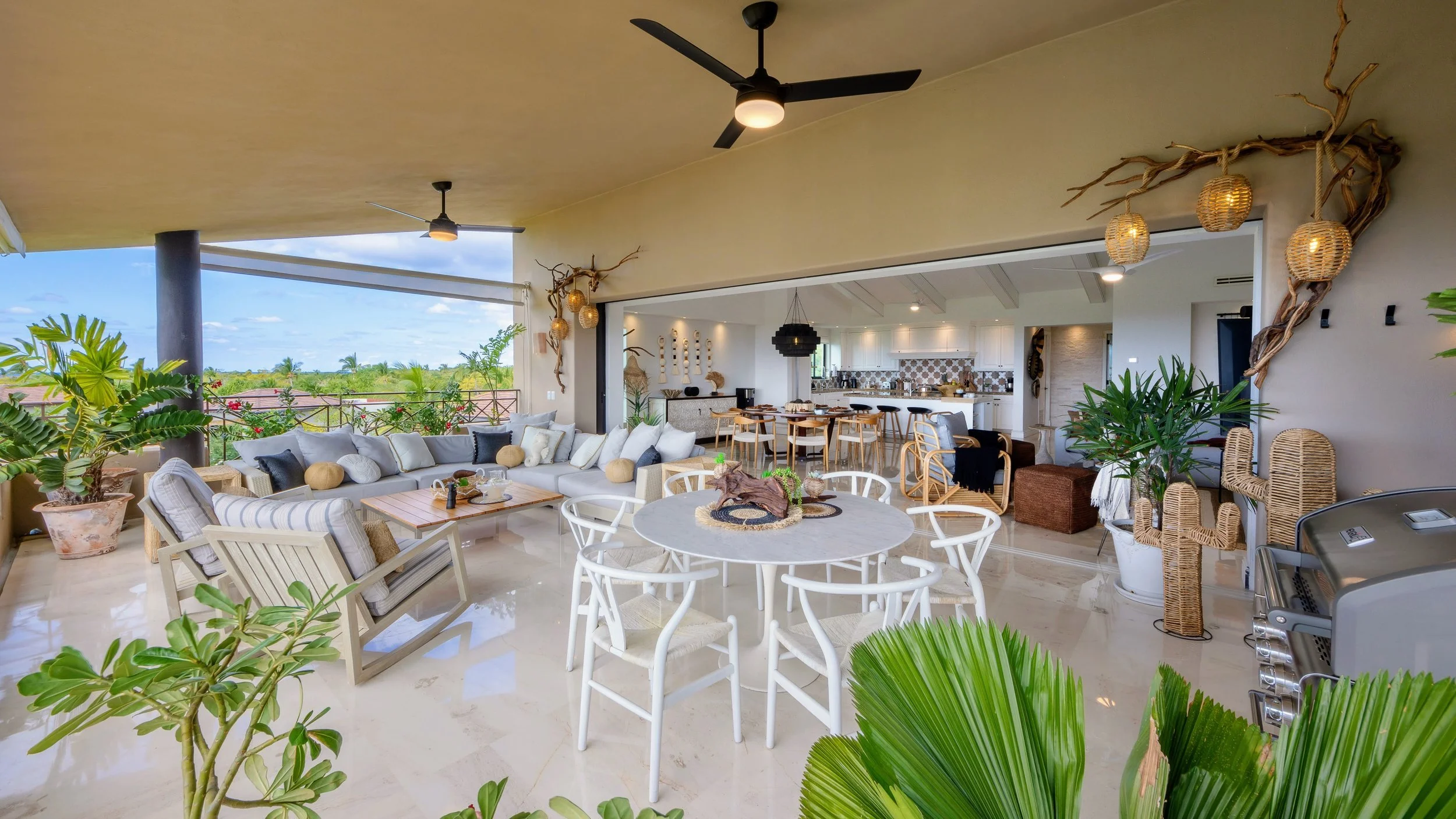 Open-concept tropical outdoor balcony with seating, dining area, kitchen, and lush green plants.