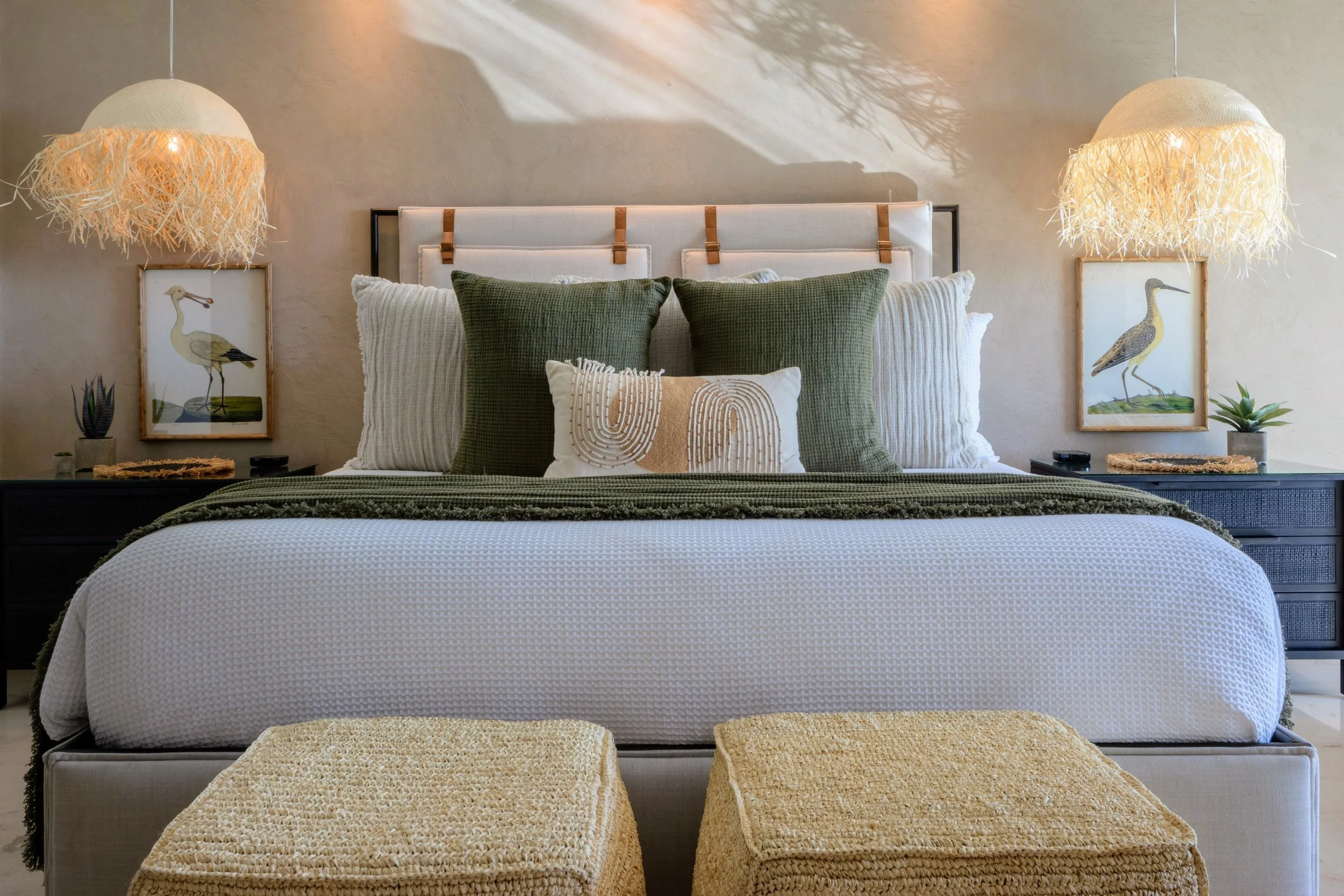 A neatly made bed with green, white, and beige decorative pillows, flanked by matching side tables with framed bird artwork, potted plants, and hanging woven light fixtures in a modern bedroom.