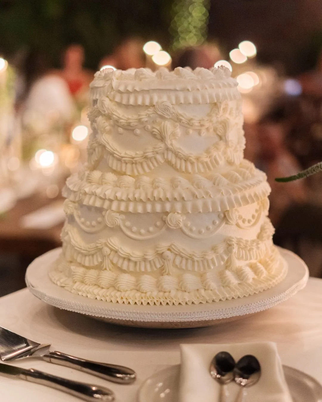 A tall, multi-tiered white wedding cake decorated with ornate piping and intricate designs, set on a cake stand with a dessert plate and utensils in front. Blurred background with warm lights and guests.