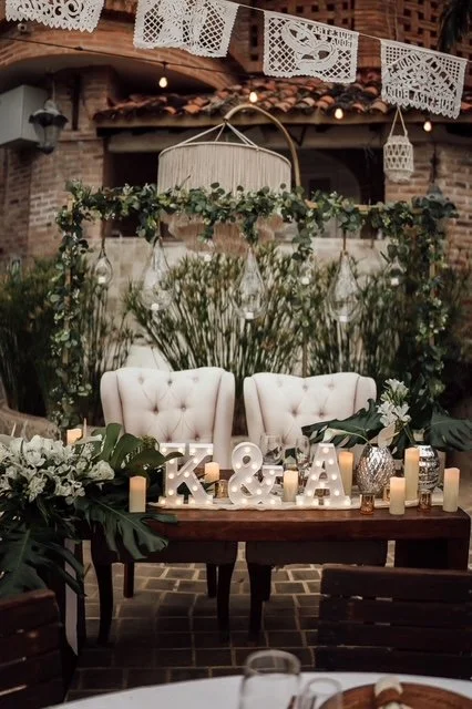 Decorated outdoor patio with white tufted chairs, greenery, string lights, paper lanterns, and large decorative letters 'T K & A' on a wooden table.