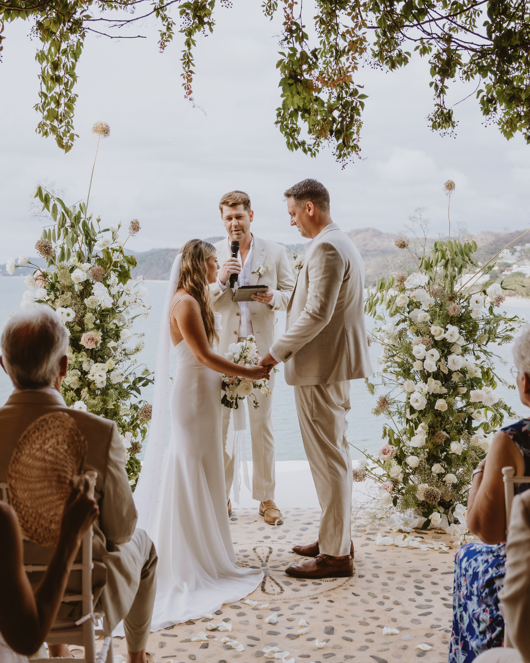 A wedding ceremony taking place outdoors with the ocean and hills in the background. The bride and groom are holding hands, exchanging vows, and standing in front of a floral arch with white and pink flowers. An officiant is holding a microphone and reading from a book, while guests seated on white chairs watch the ceremony.