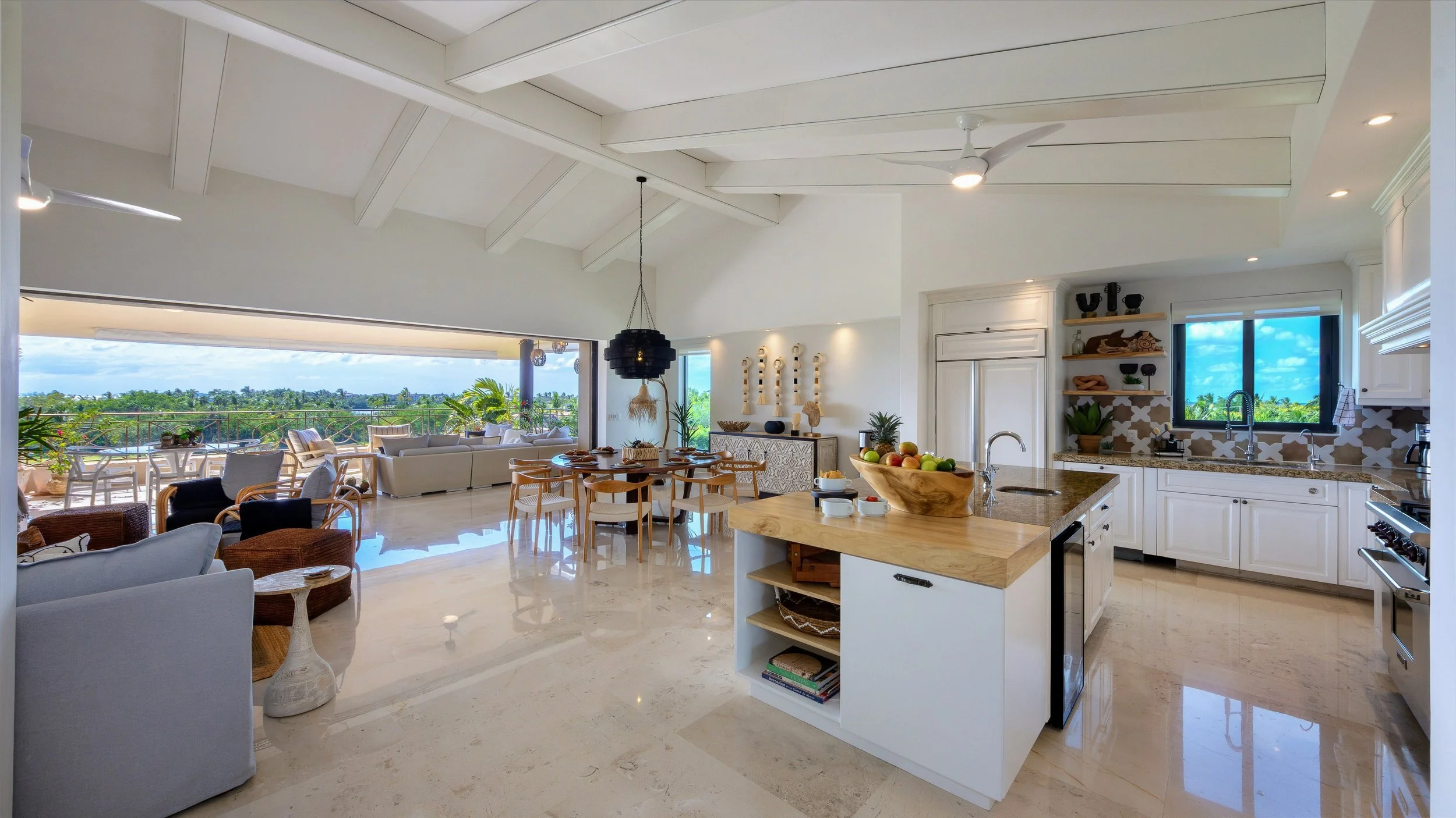 Open-concept living room and kitchen with white walls and high vaulted ceiling, large windows revealing a lush green landscape, and modern furnishings including a beige sectional sofa, wooden chairs, and a kitchen island with a bowl of fruit.